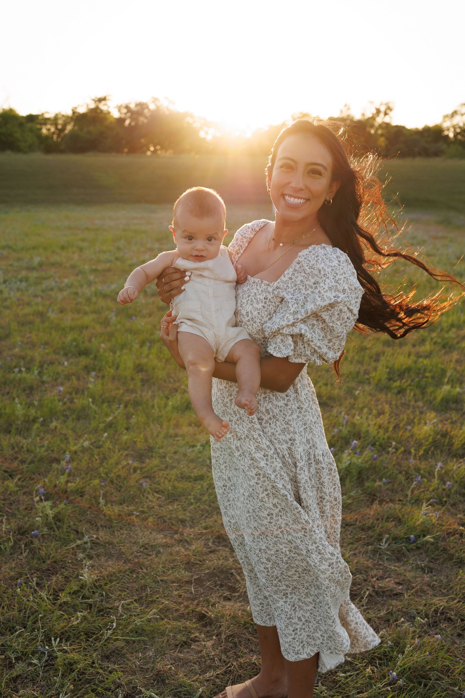 Sun-drenched portrait of parent and baby in floral dress standing in golden field at sunset.