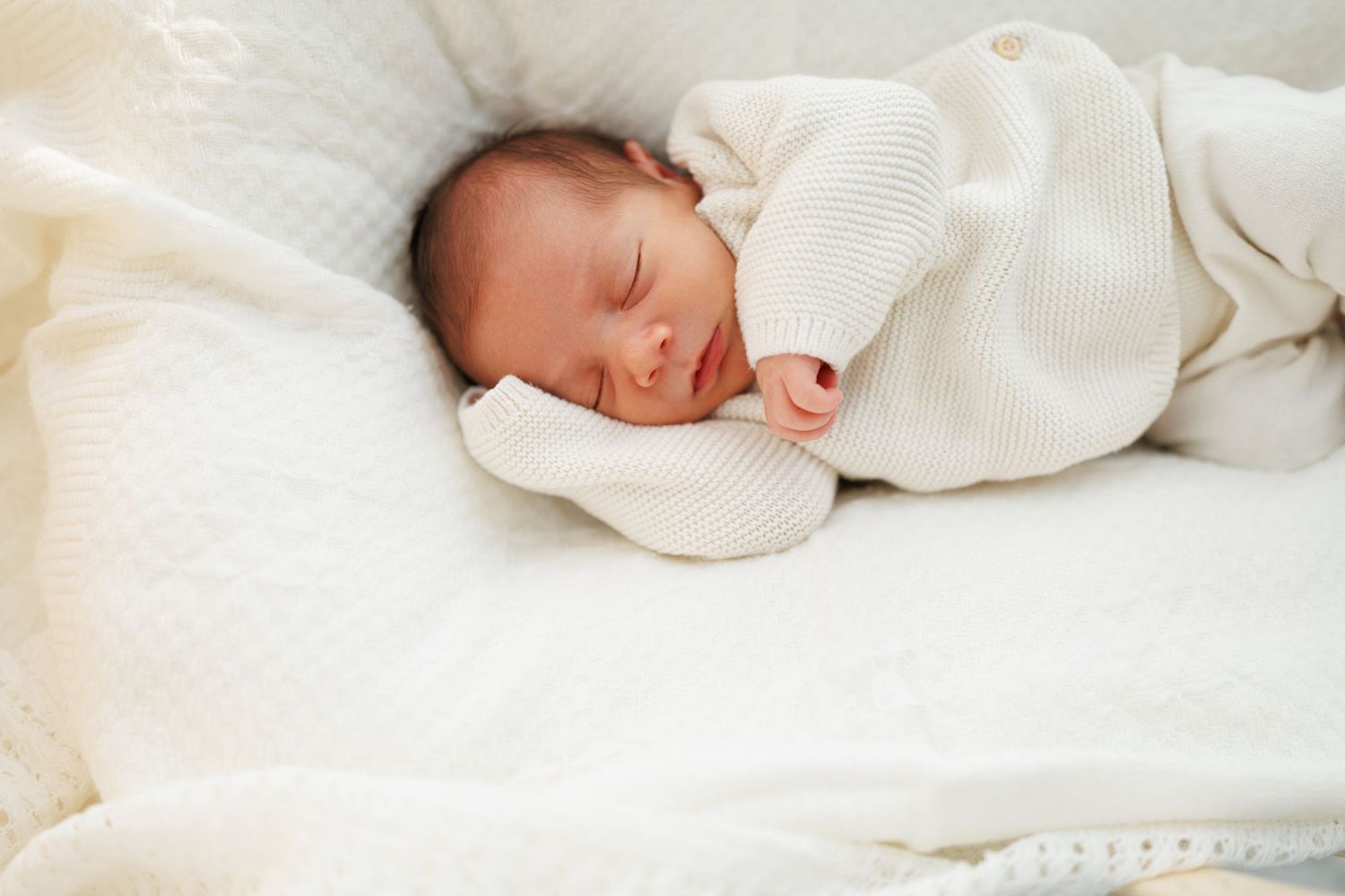 A newborn baby sleeps peacefully swaddled in white blankets on a soft white surface.