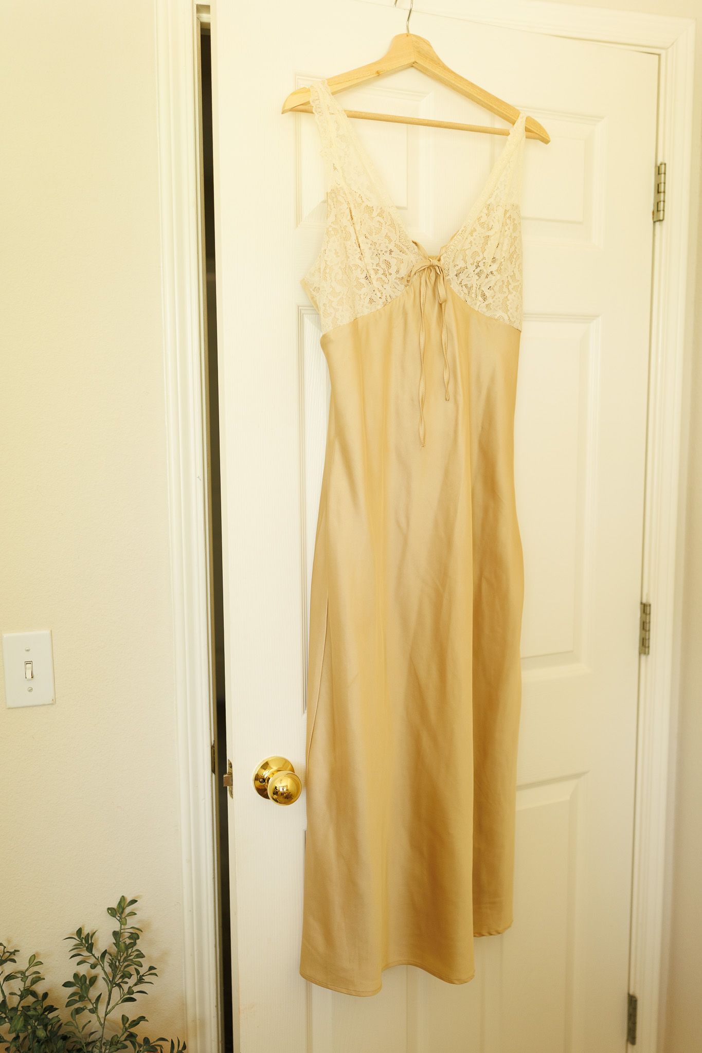 A cream-colored satin nightgown with lace detail hangs on a wooden hanger against a white door.