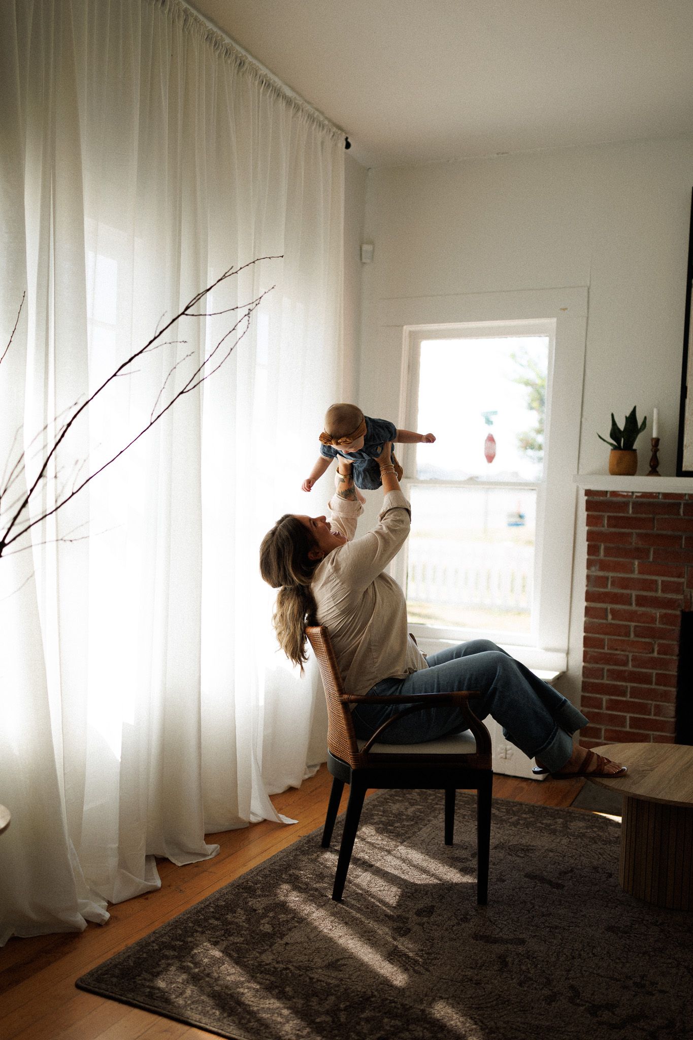 Mother lifting smiling baby in the air while seated in a chair by curtains.