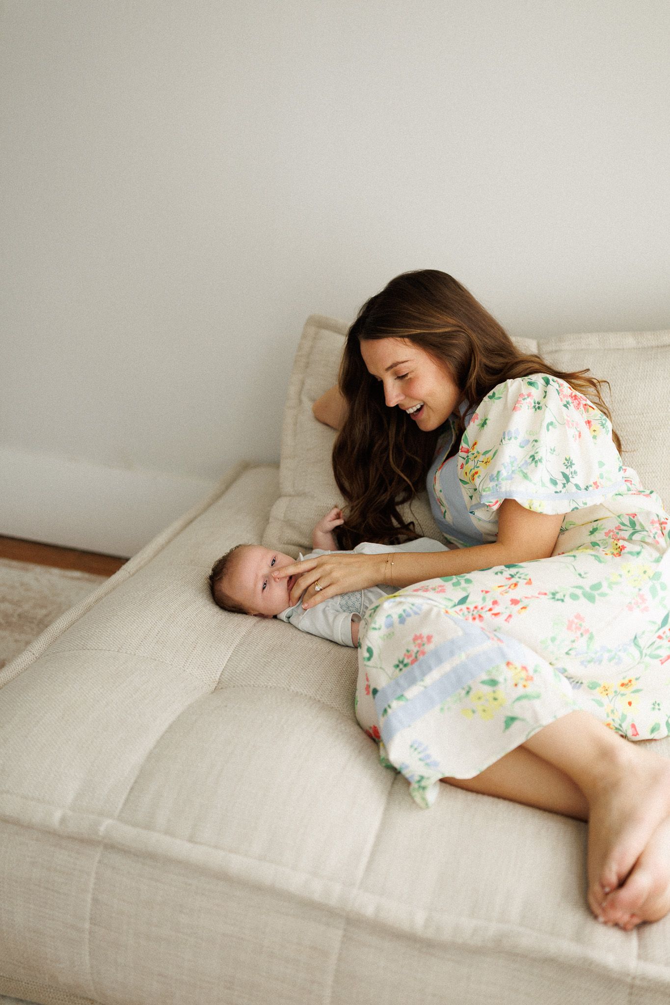 Smiling mother in floral dress leaning over newborn baby lying on beige sofa.