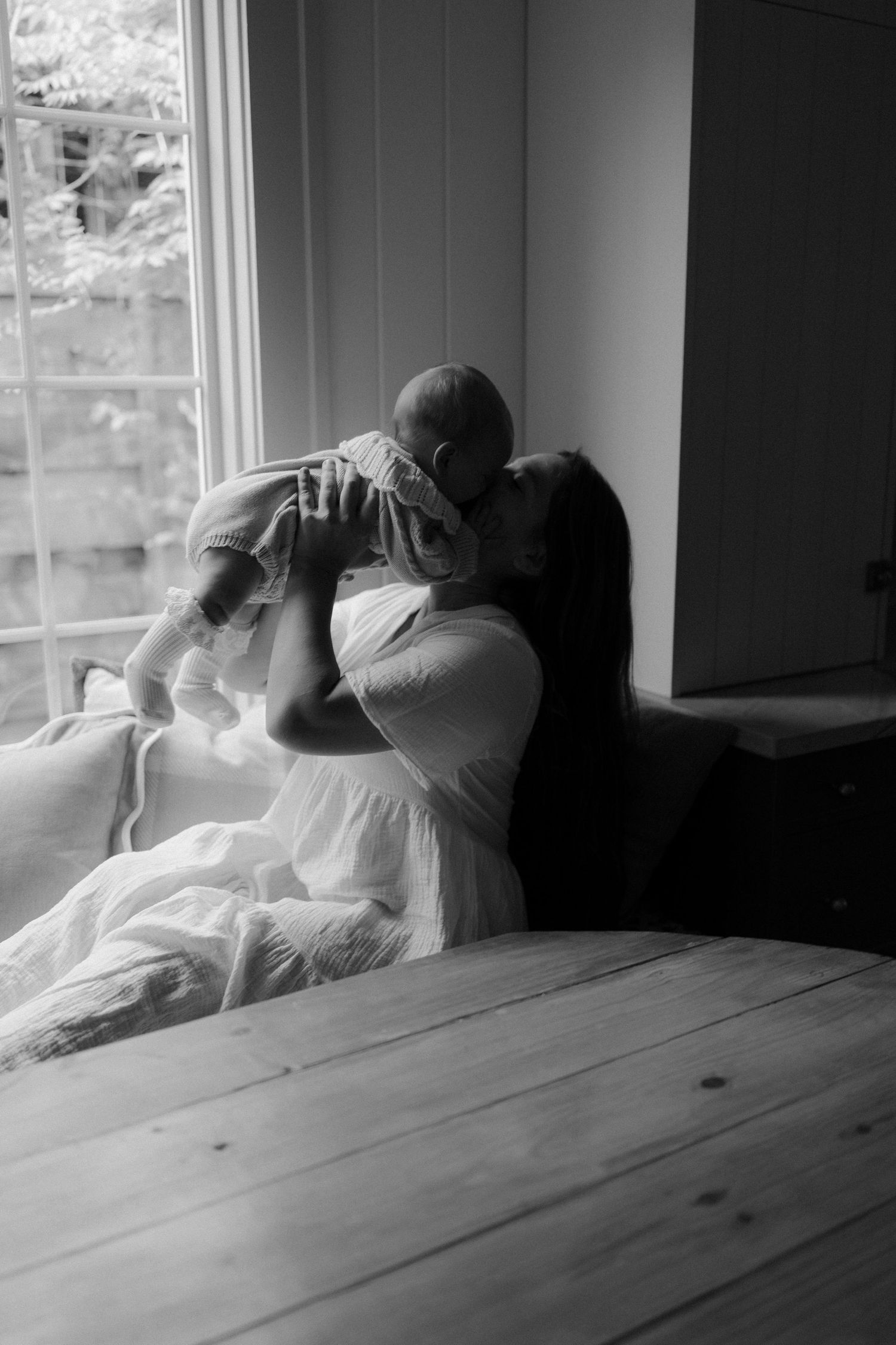 Black and white photo of a mother lifting her baby up for a kiss near a bright window indoors.