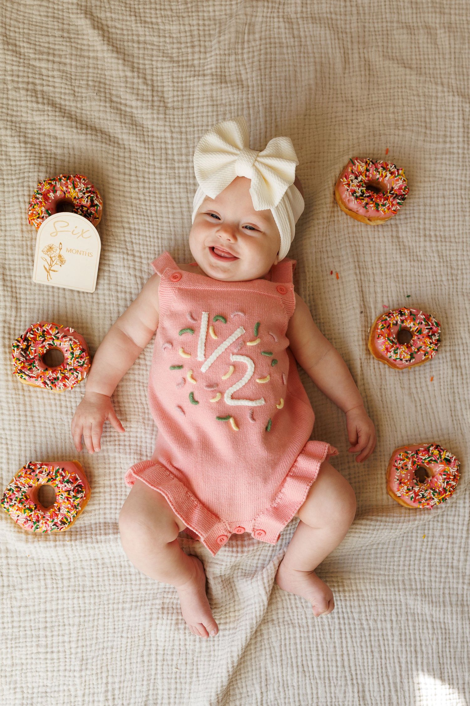 Baby wearing pink outfit with white bow lying on cream blanket surrounded by chocolate frosted donuts.
