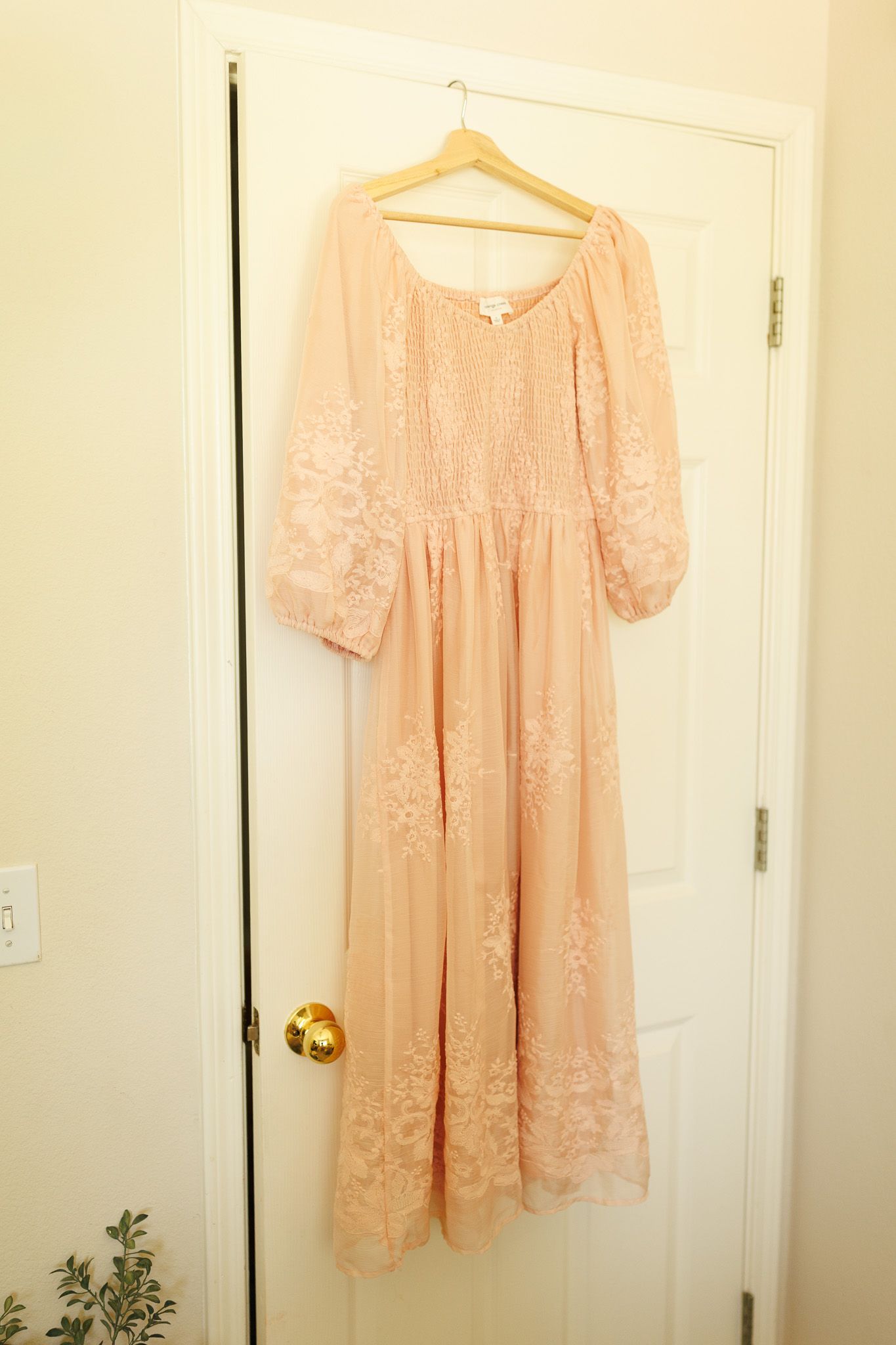 A vintage-style peach lace maxi dress with long sleeves hangs on a white wooden hanger against a cream-colored door.