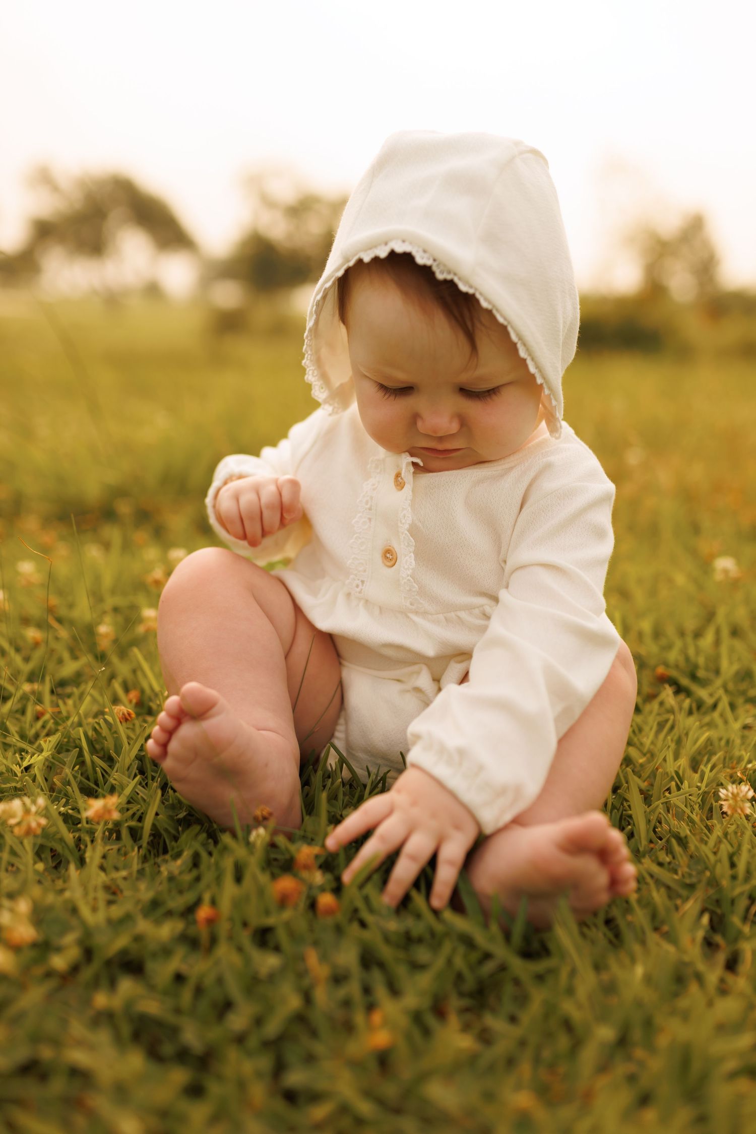 Small child wearing white bonnet and dress sits on grassy field examining yellow wildflowers in warm sunlight.