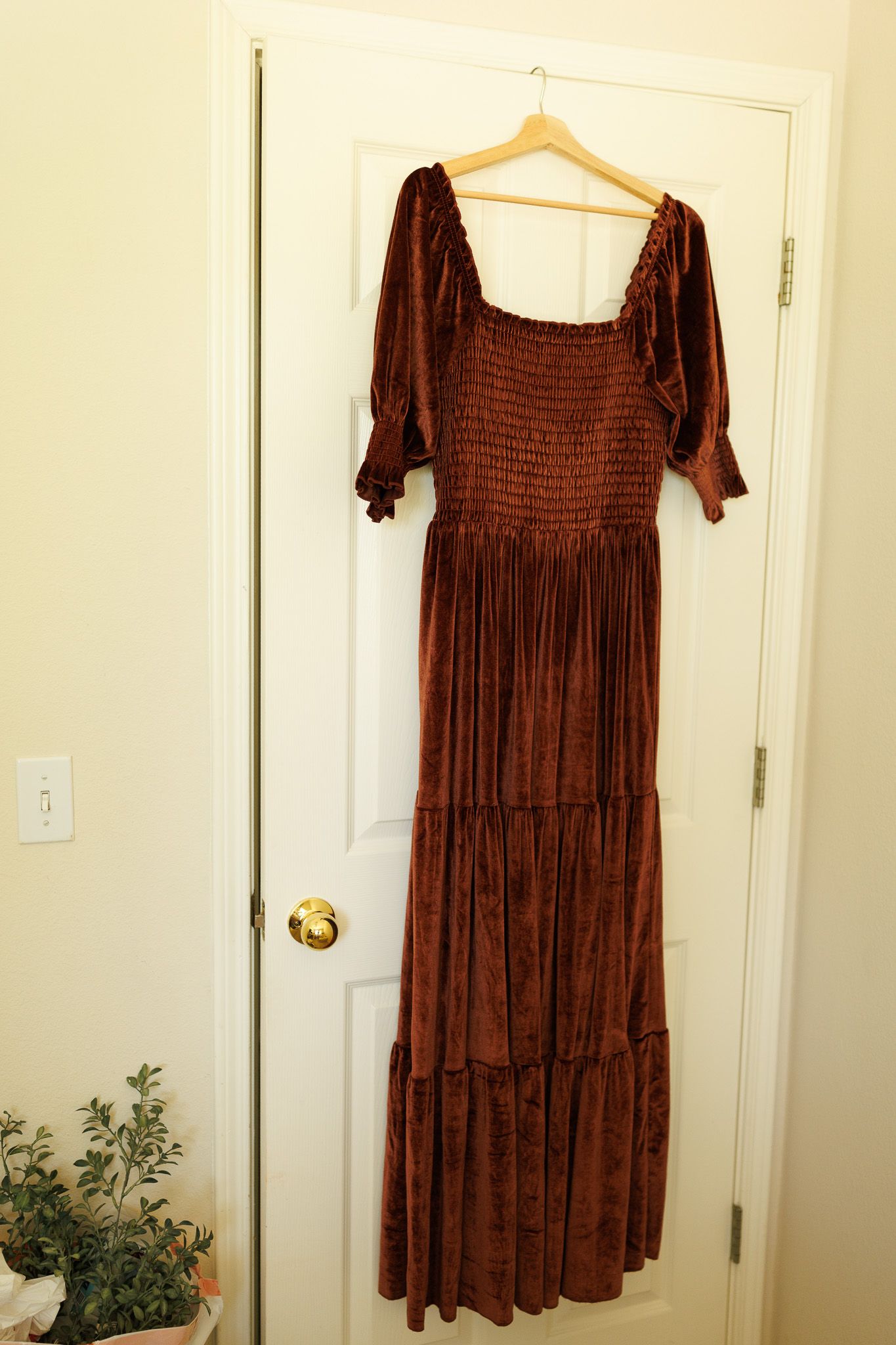 A long brown velvet maxi dress with smocked bodice and tiered skirt hangs on a wooden hanger against a white door.