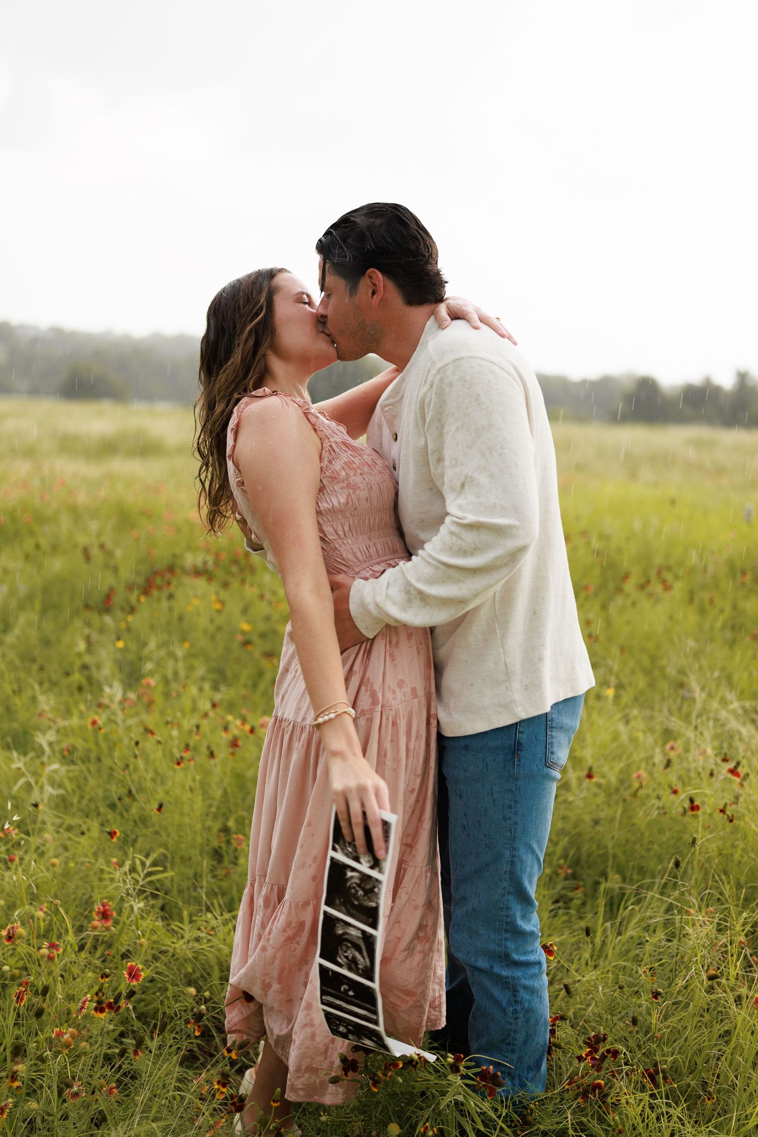 Couple kissing in a wildflower field, woman in pink dress holding ultrasound photo, romantic pregnancy announcement outdoors.