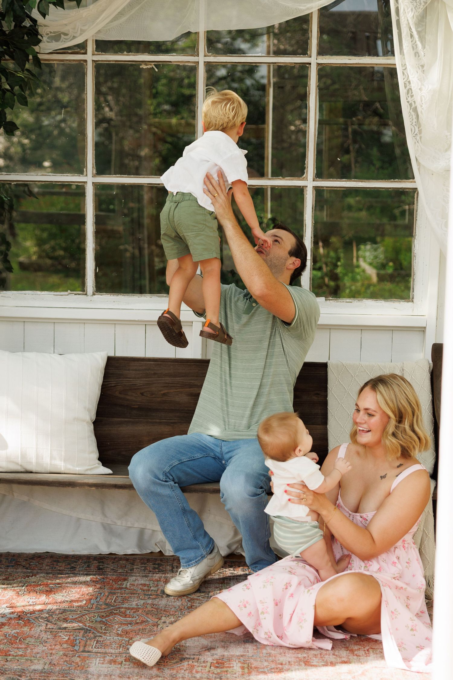 A happy family enjoys playtime outdoors, with dad lifting toddler while mom holds baby on a cozy porch.
