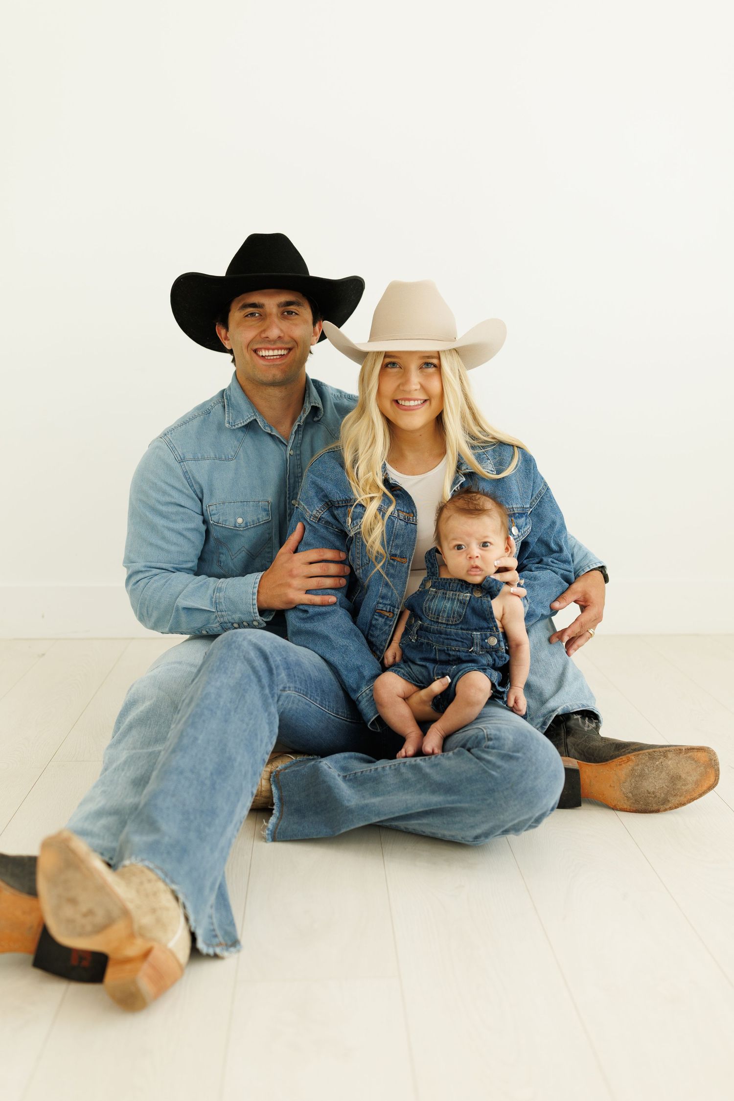 A family in matching denim outfits and cowboy hats sits together on a white backdrop for a casual portrait session.