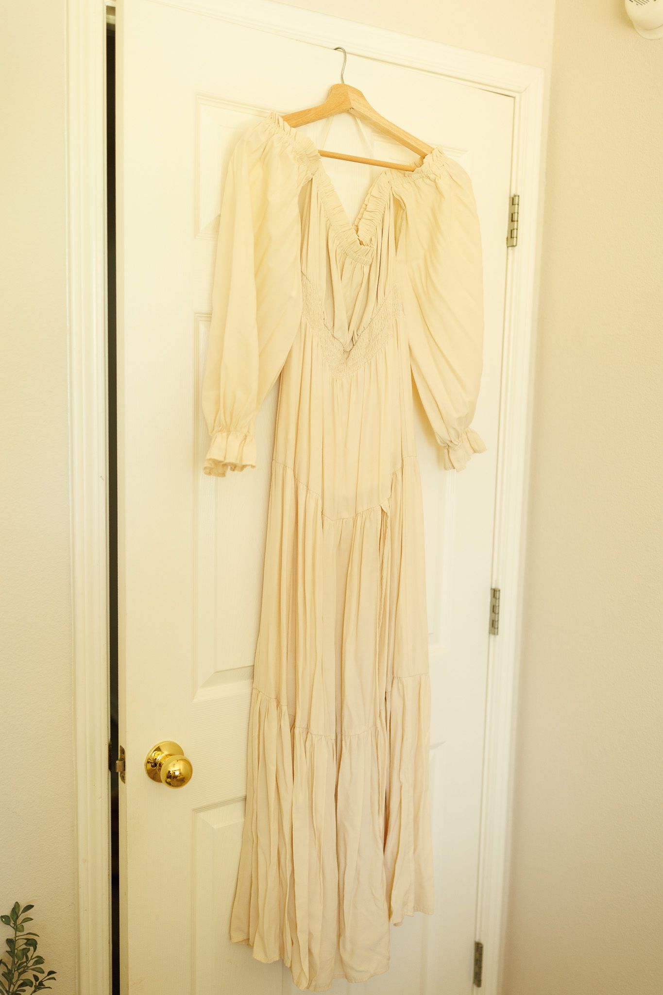 A cream colored vintage-style dress with long sleeves hangs on a white door against a light wall.