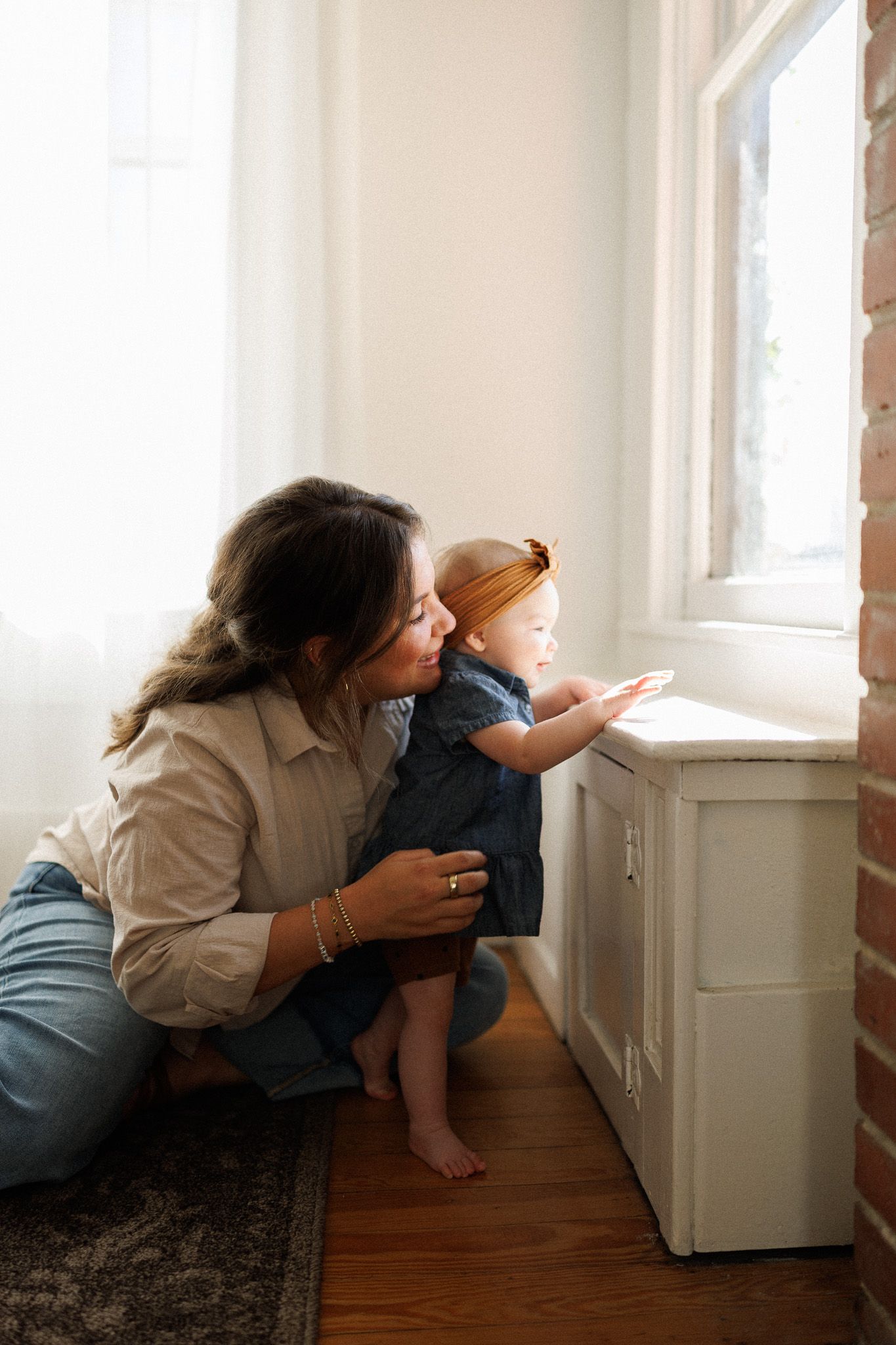 A mother crouches beside her baby girl in a blue dress and orange headband, both gazing out a sunlit window together.