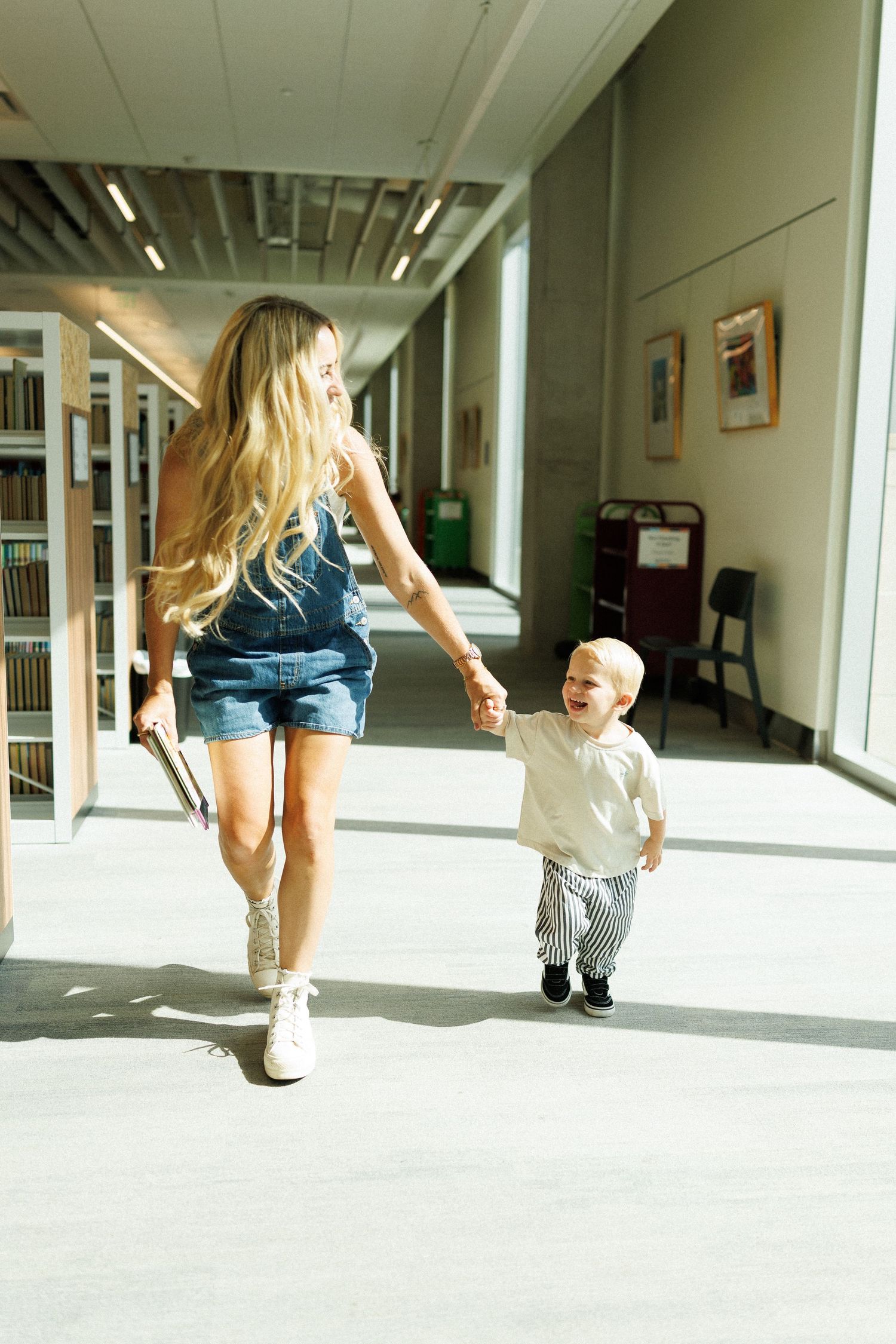 A smiling woman in denim overalls walks hand-in-hand with a laughing toddler through a bright library hallway.