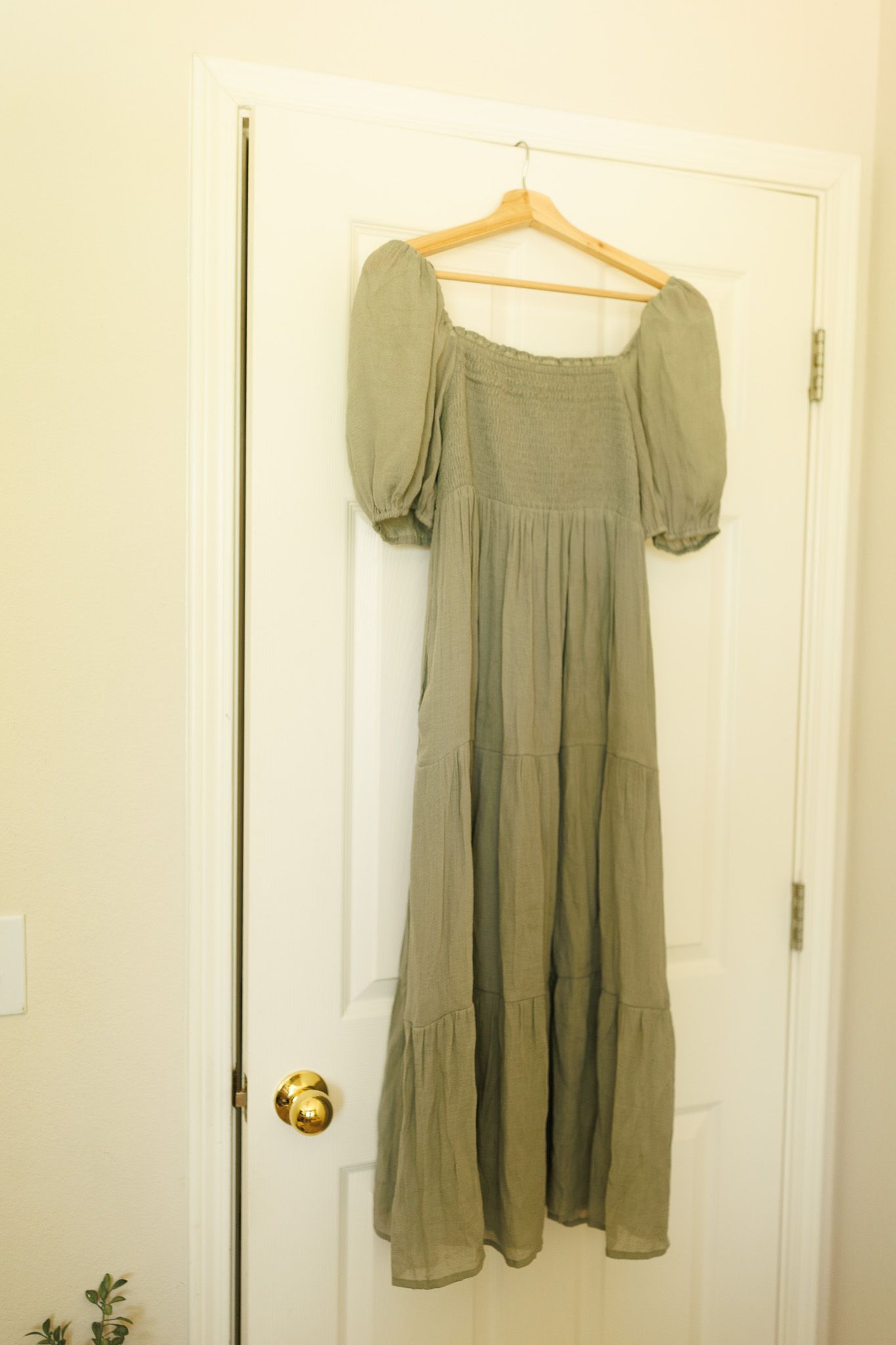Olive green tiered maxi dress with puff sleeves hangs on wooden hanger against white door.