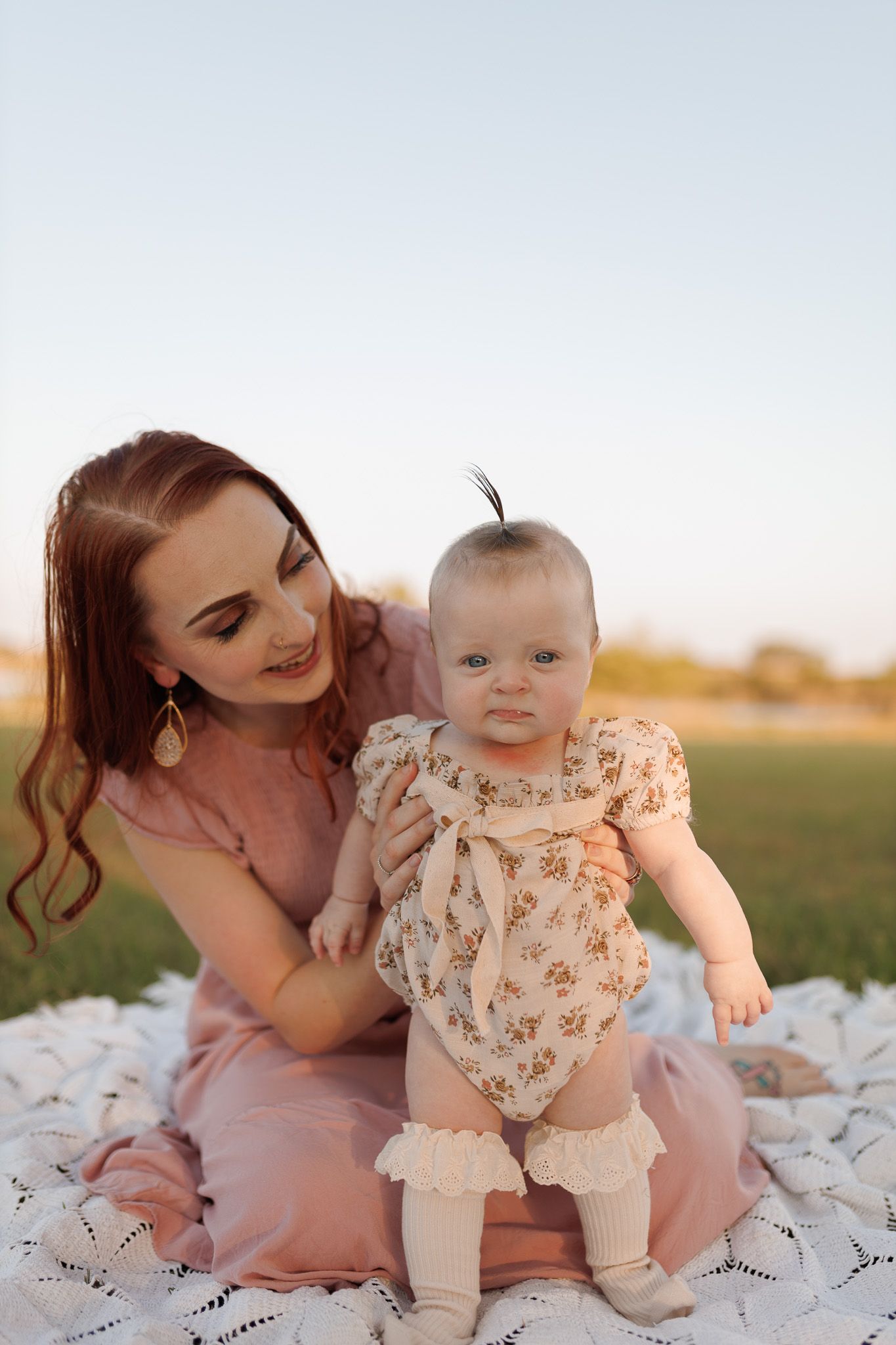 Parent and baby share tender outdoor moments while sitting on white blanket in grassy field at sunset.