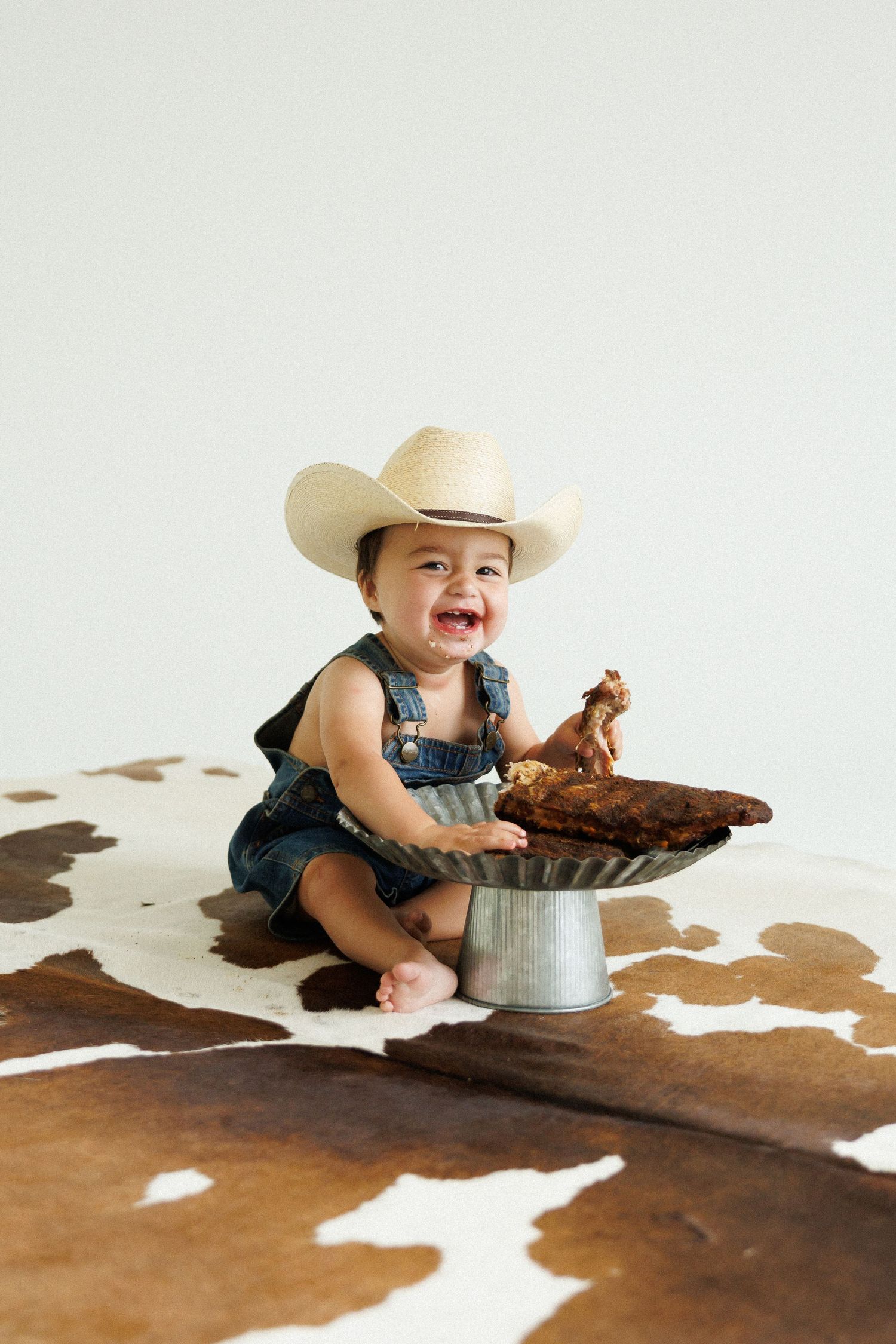 Smiling baby in denim overalls and cowboy hat sitting on cowhide rug with smashed cake on metal stand.