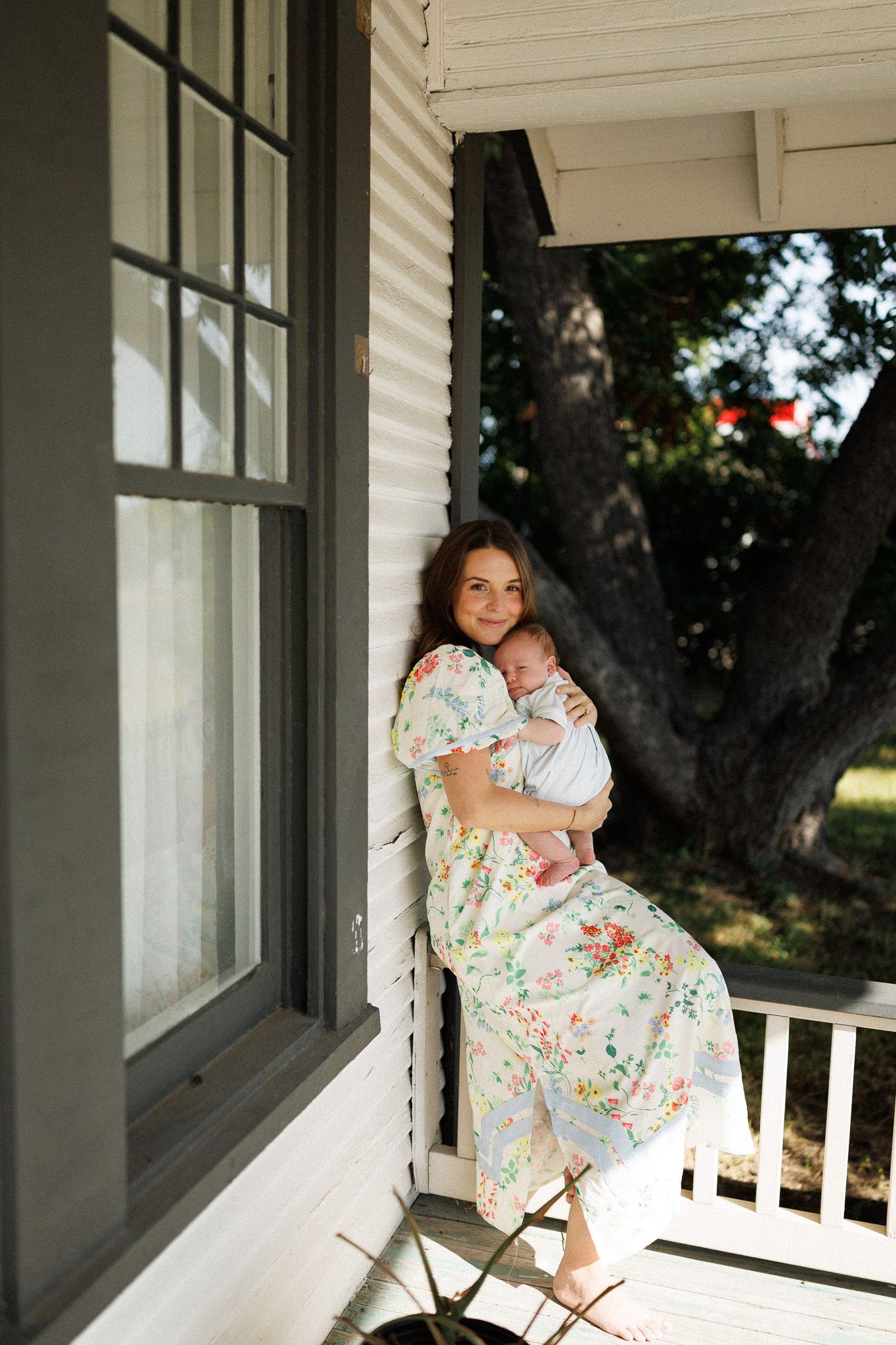 Mother in floral dress holding newborn baby on a white farmhouse porch.