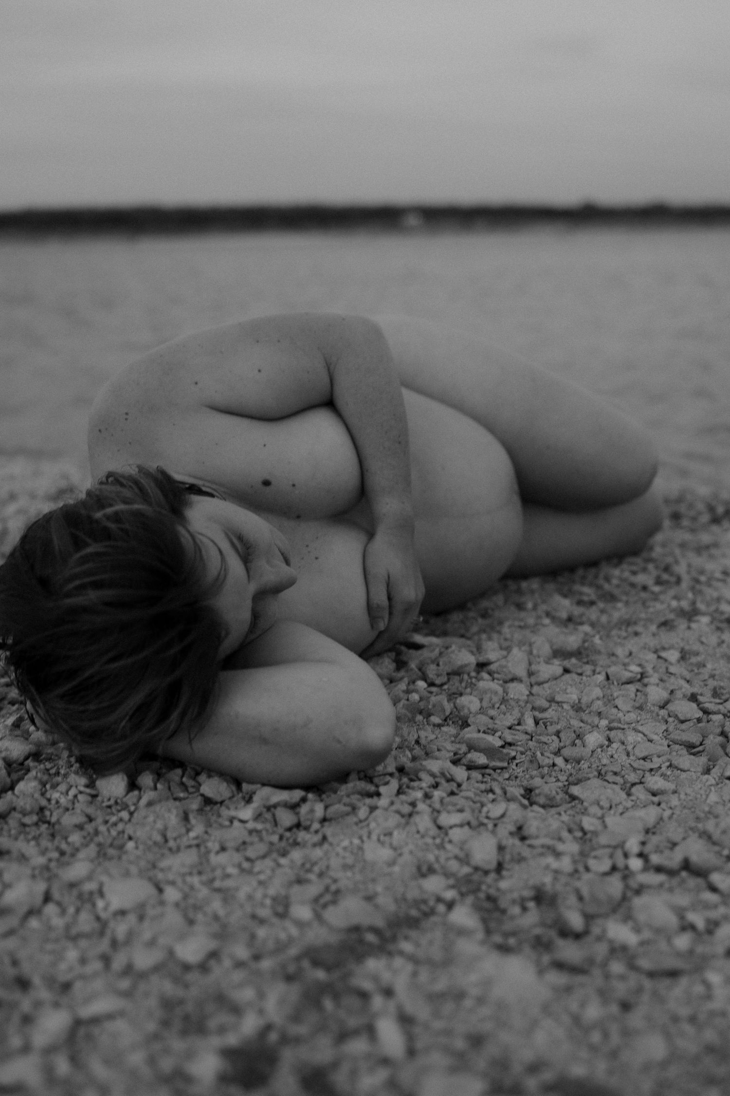 Artistic black and white photograph of a nude figure curled up on a rocky shore in a fetal position.