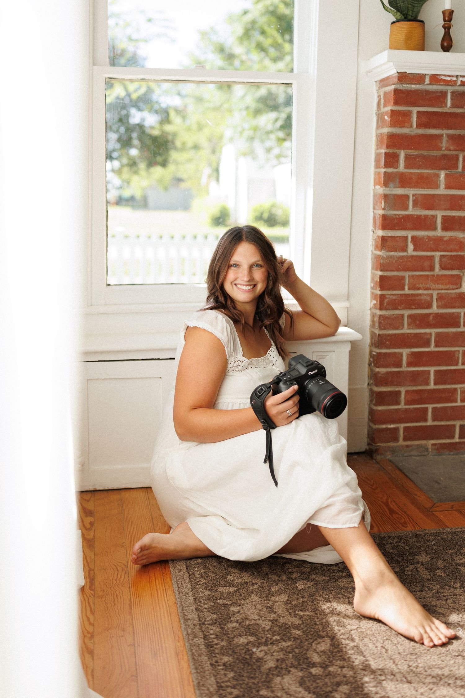 Smiling woman in white dress sitting on hardwood floor holding DSLR camera near bright window and brick fireplace.
