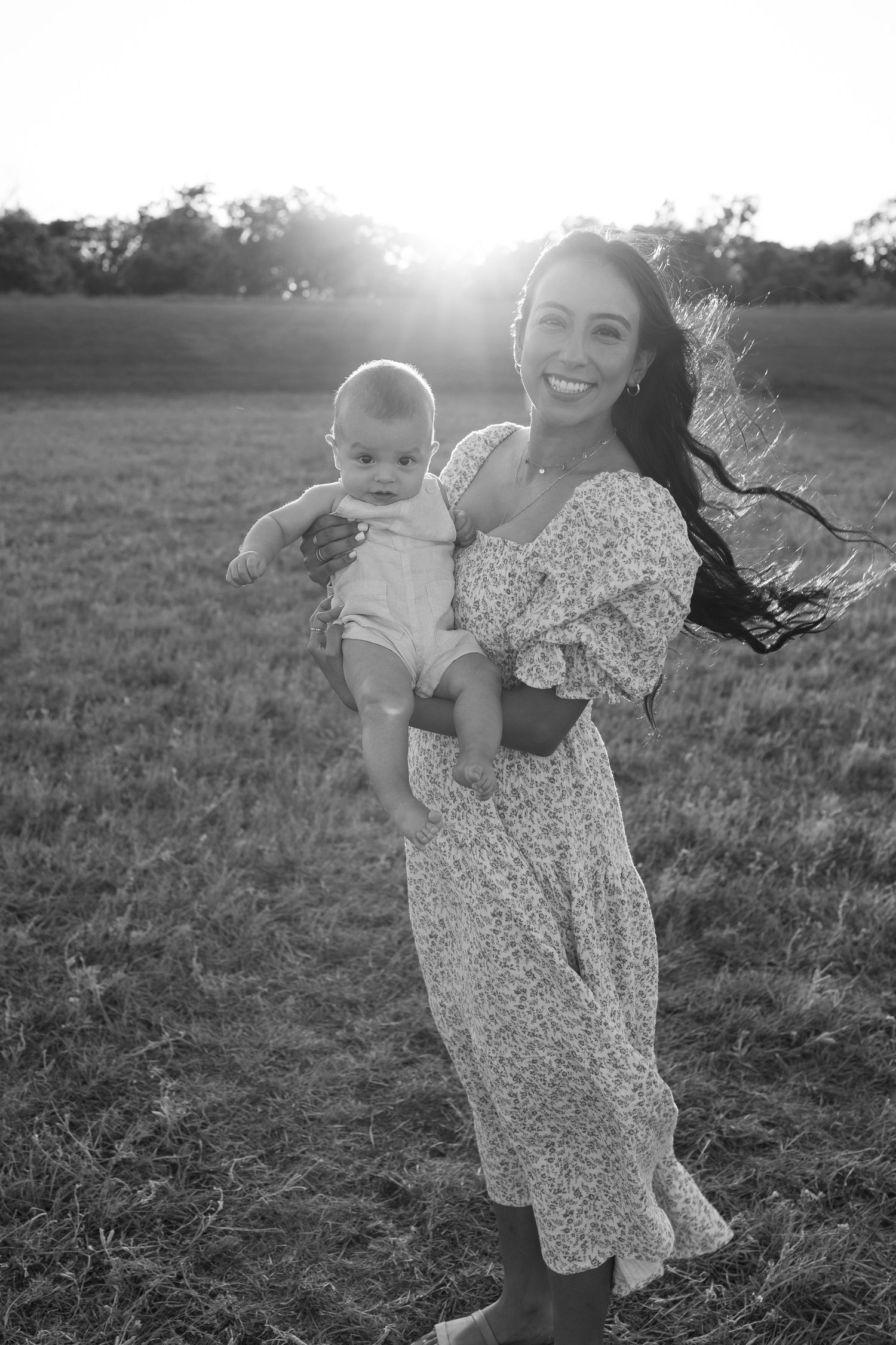 A tender black and white portrait of a mother and baby in a floral dress standing in a sunlit field at dusk.