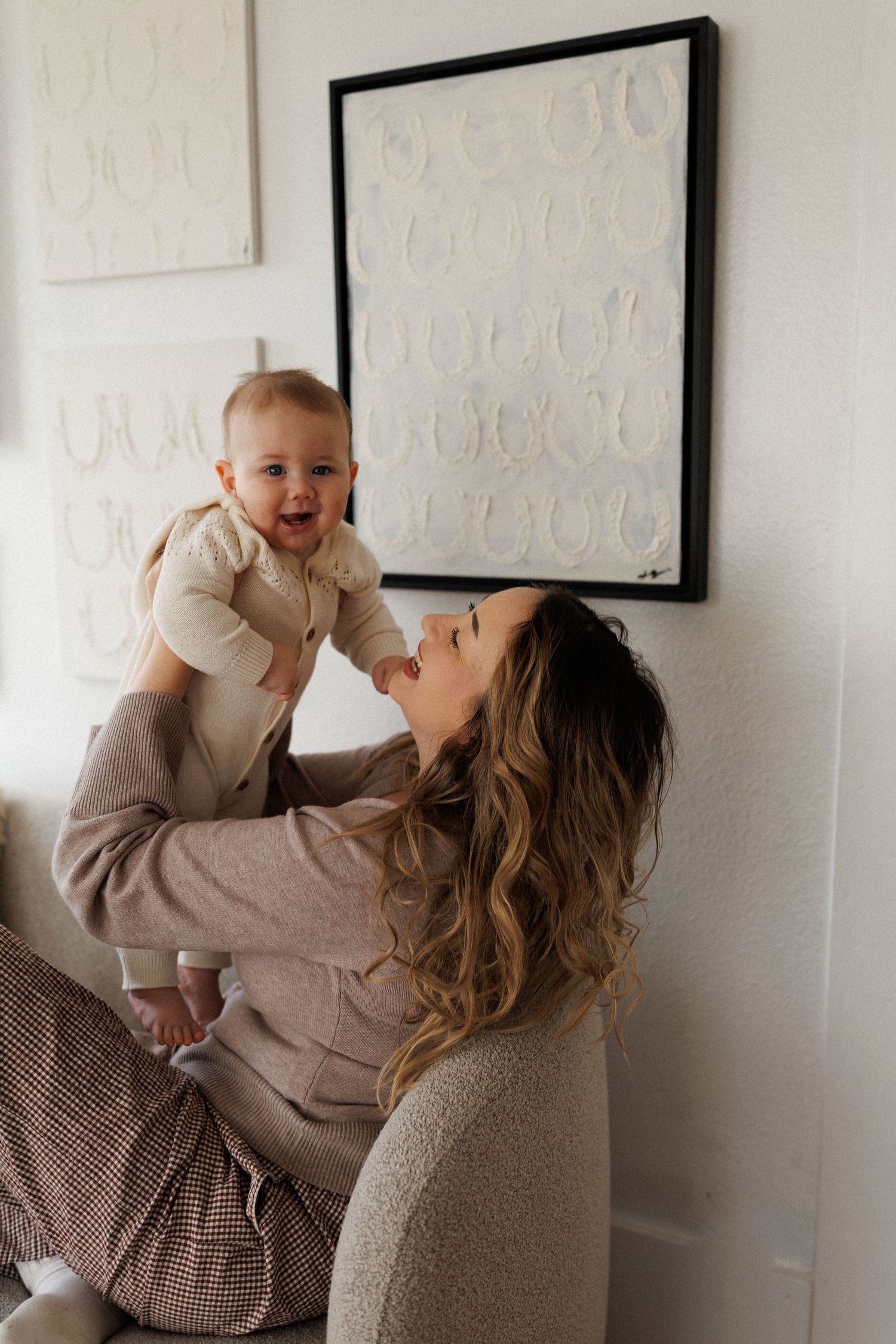 A parent in a beige sweater lifts and plays with a baby against a white wall with framed artwork.