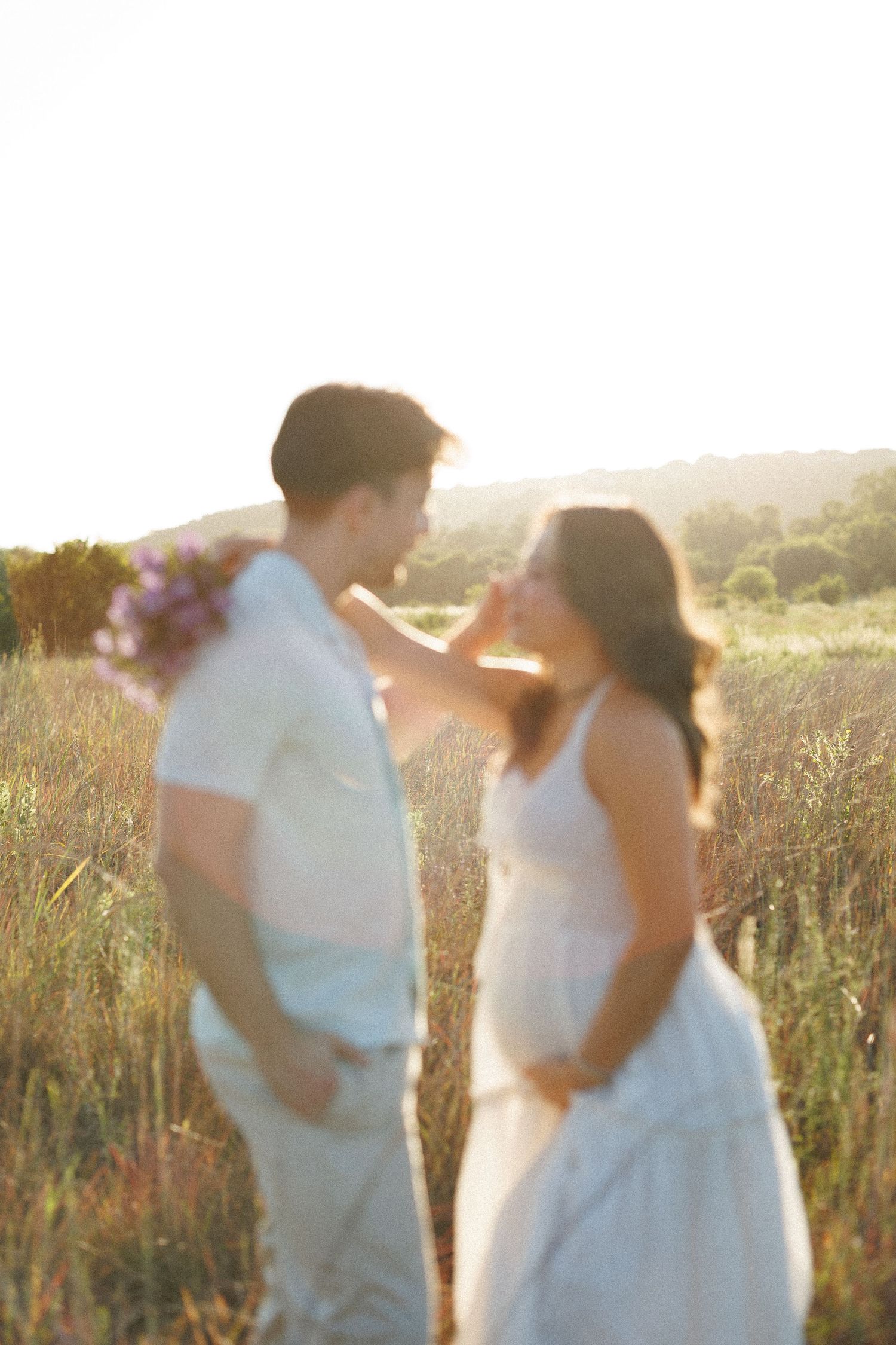 A couple shares a tender moment in a sunlit field, the woman in a white dress touching the man's face.