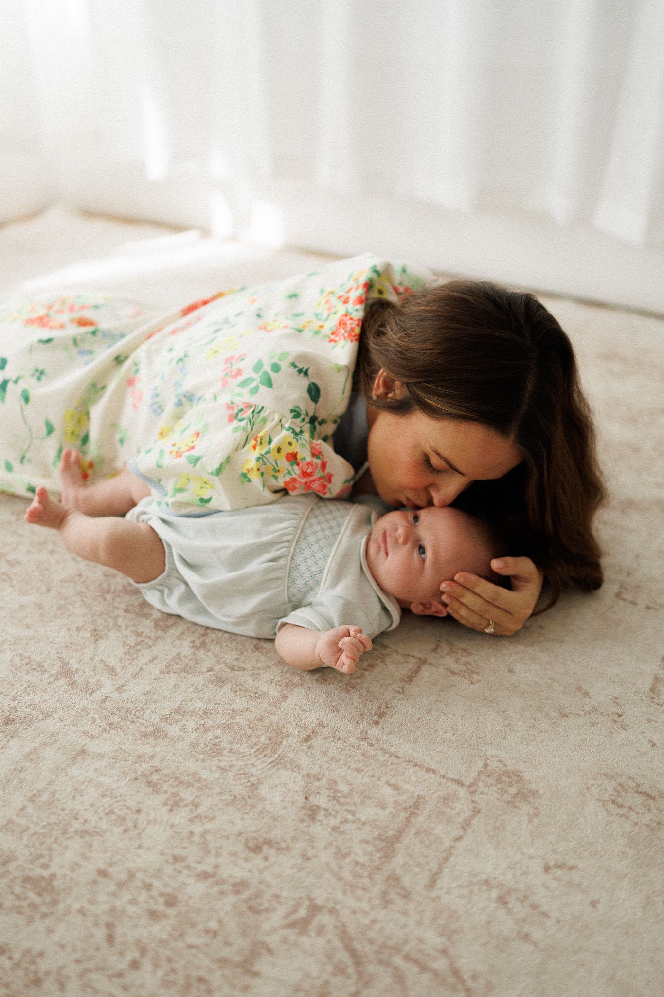 Mother in floral dress kissing newborn baby lying on carpet in sunlit room.