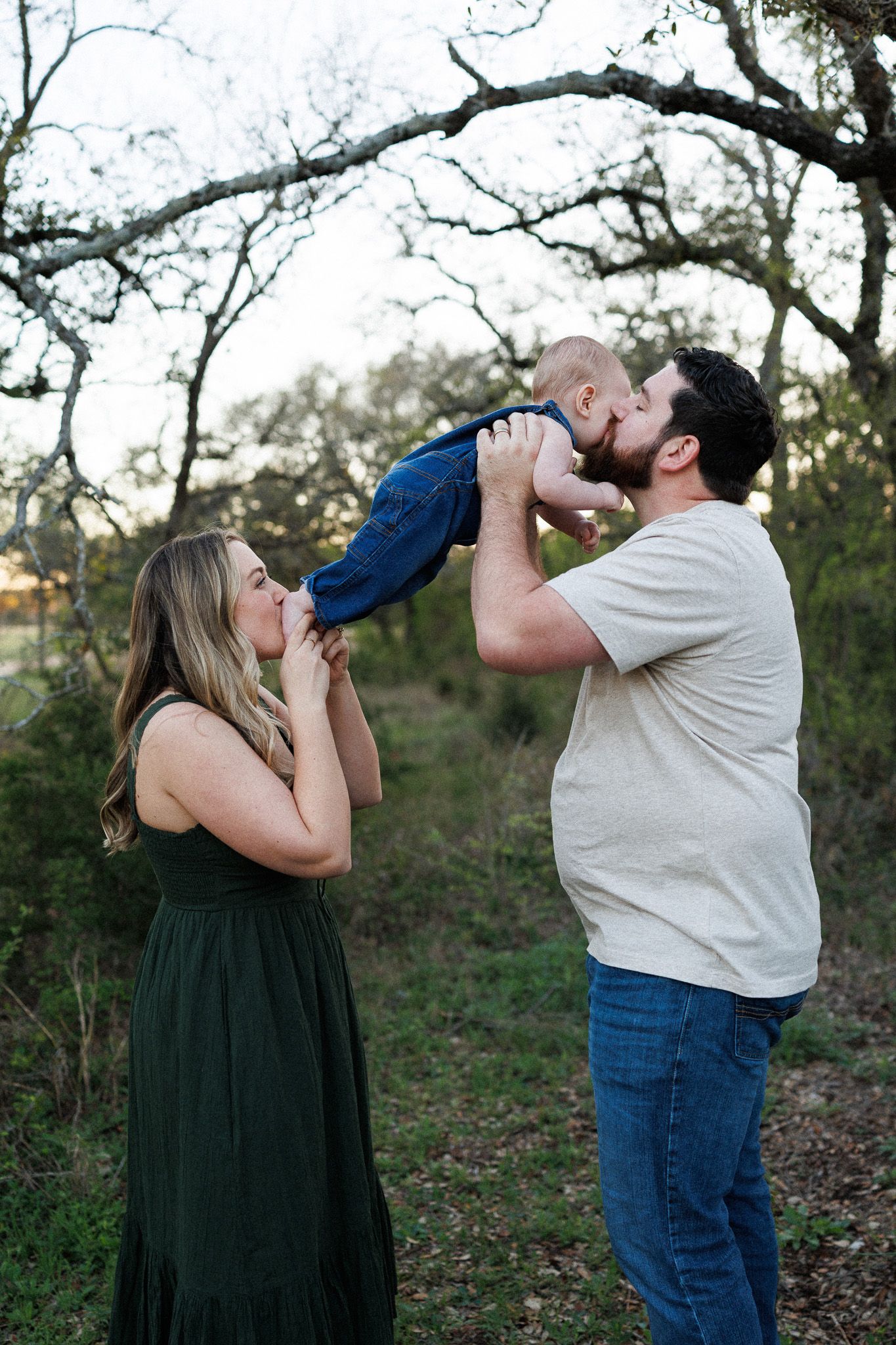 A couple in casual attire playfully drinks from wine bottles while standing in a wooded outdoor setting at dusk.