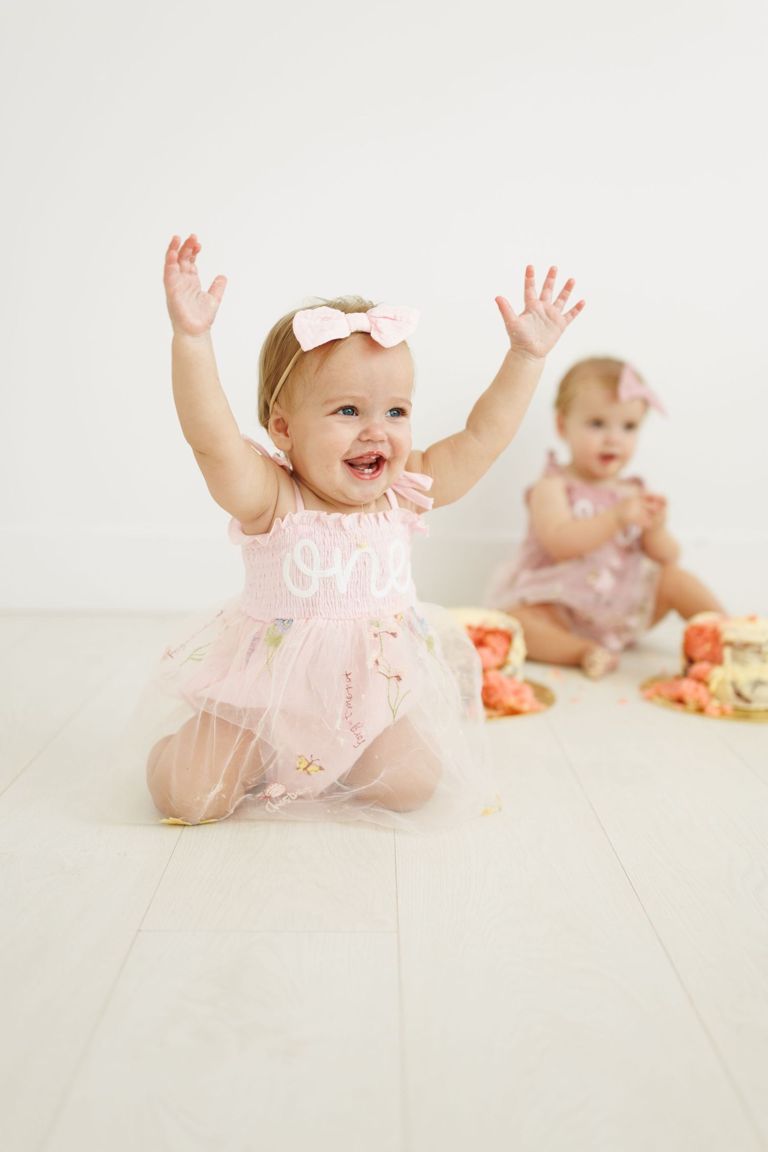 Twin baby girls in pink floral dresses celebrate their first birthday, one joyfully raising her hands during a cake smash.
