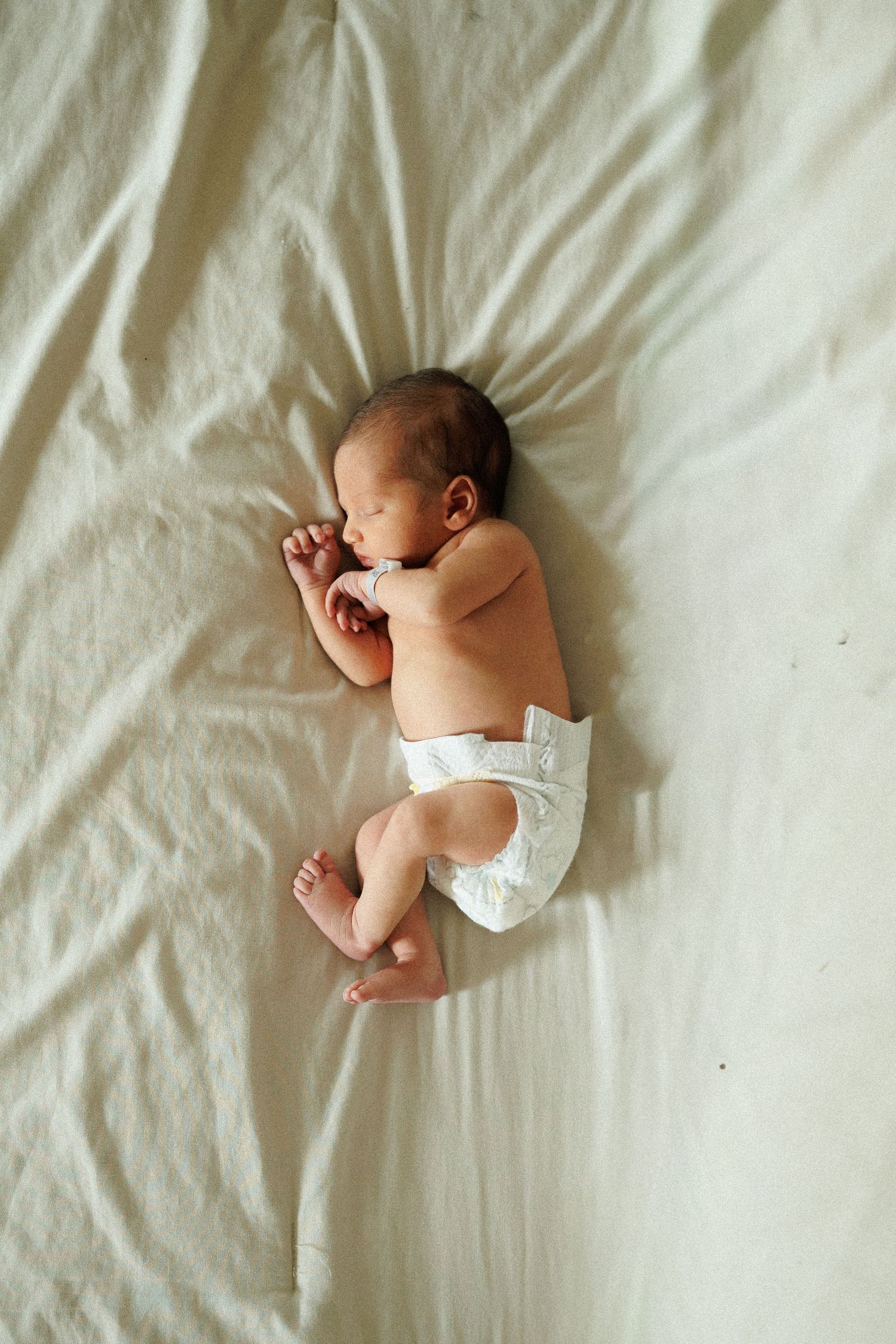 Newborn baby sleeping peacefully in a white diaper on a soft cream blanket, photographed from above.