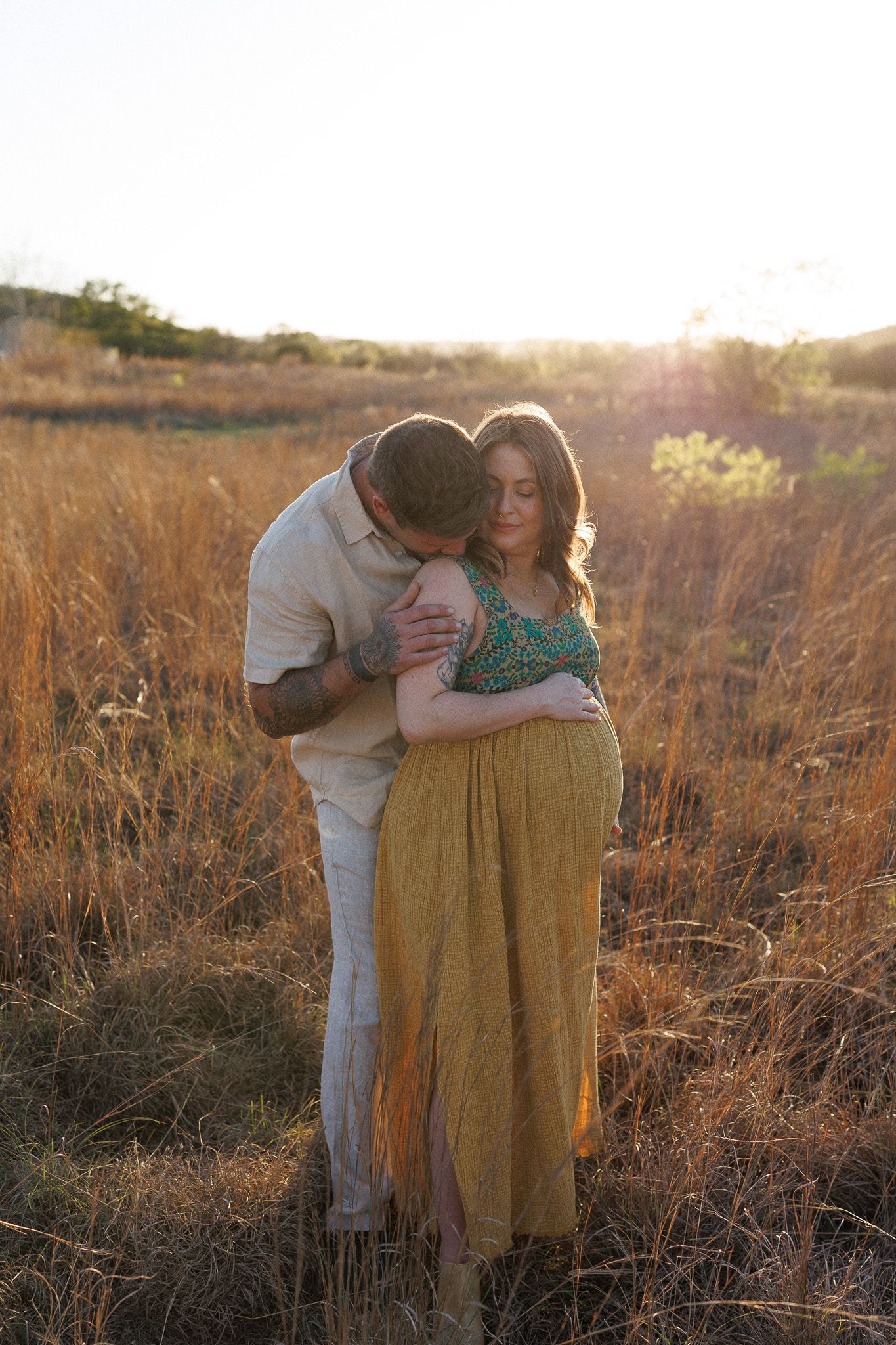 A couple embracing in a field during sunset, with the woman wearing a yellow skirt and the man wearing a light-colored shirt.