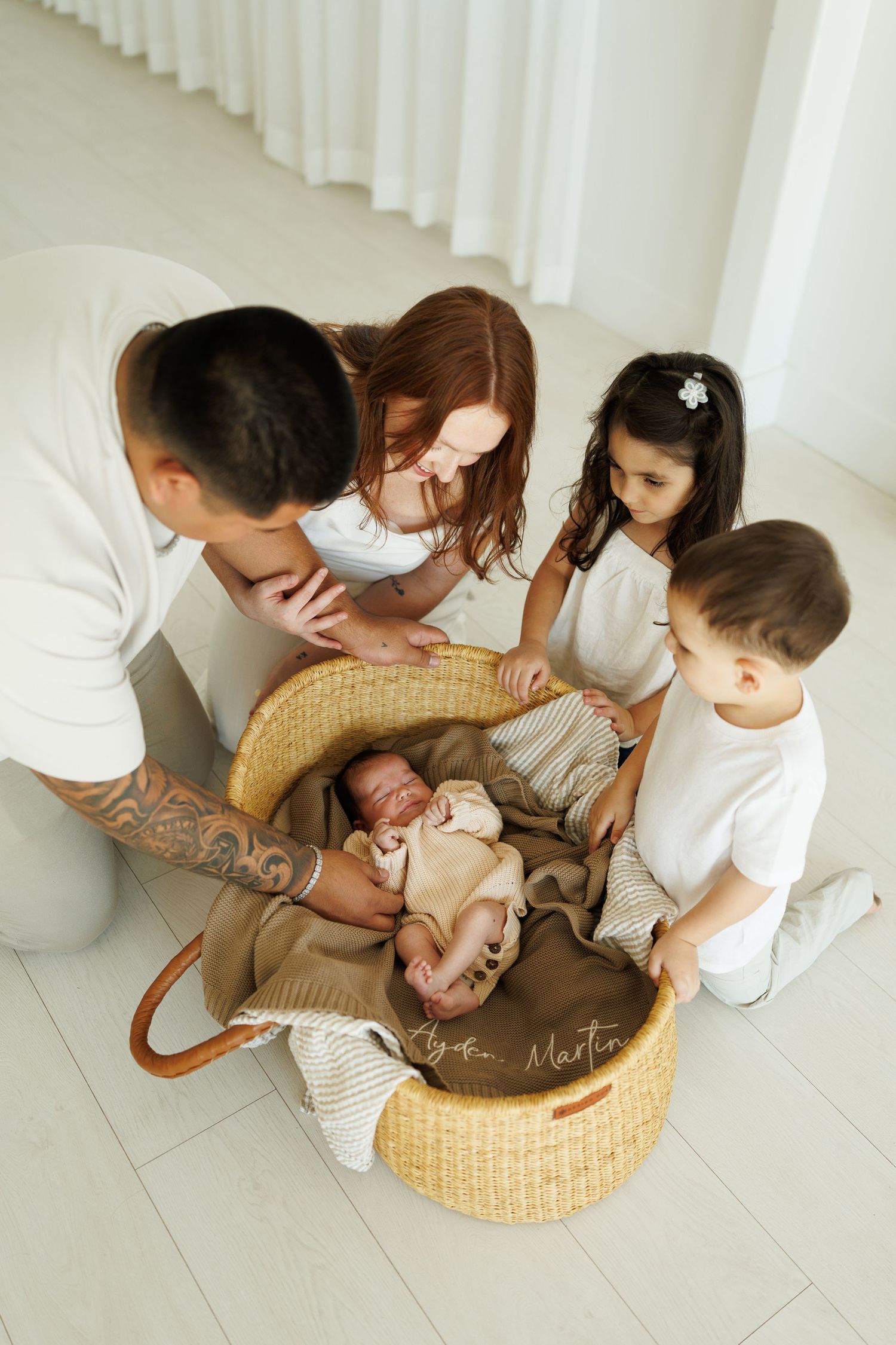 Family of five admiring newborn baby in wicker Moses basket, parents and two young children gathered together.
