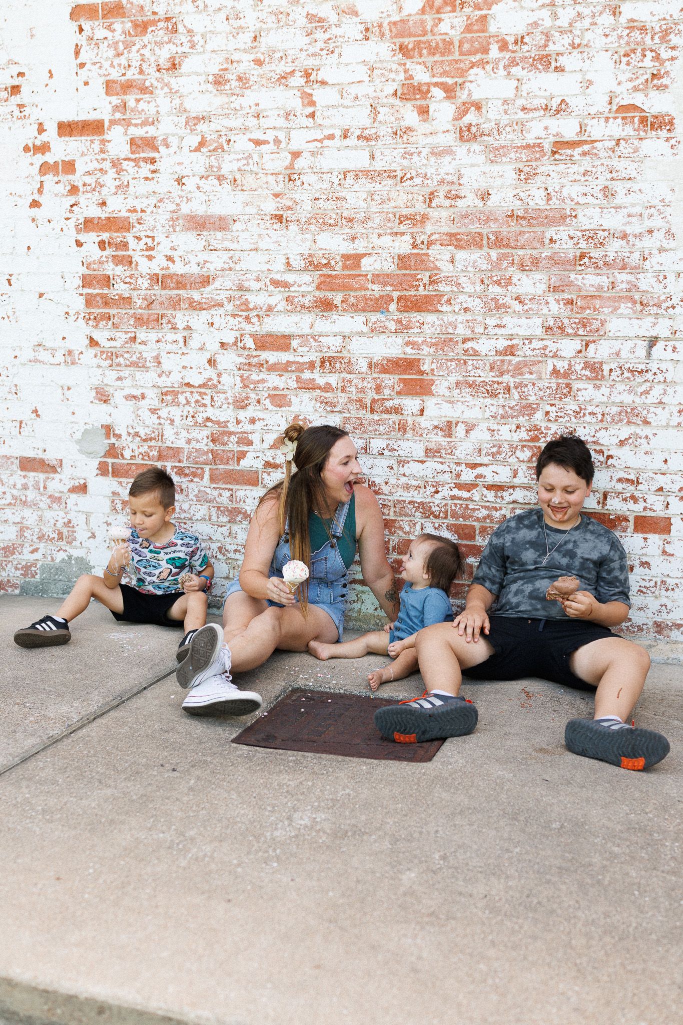 A woman and three children sit against a rustic brick wall outdoors, enjoying ice cream on a sunny day.