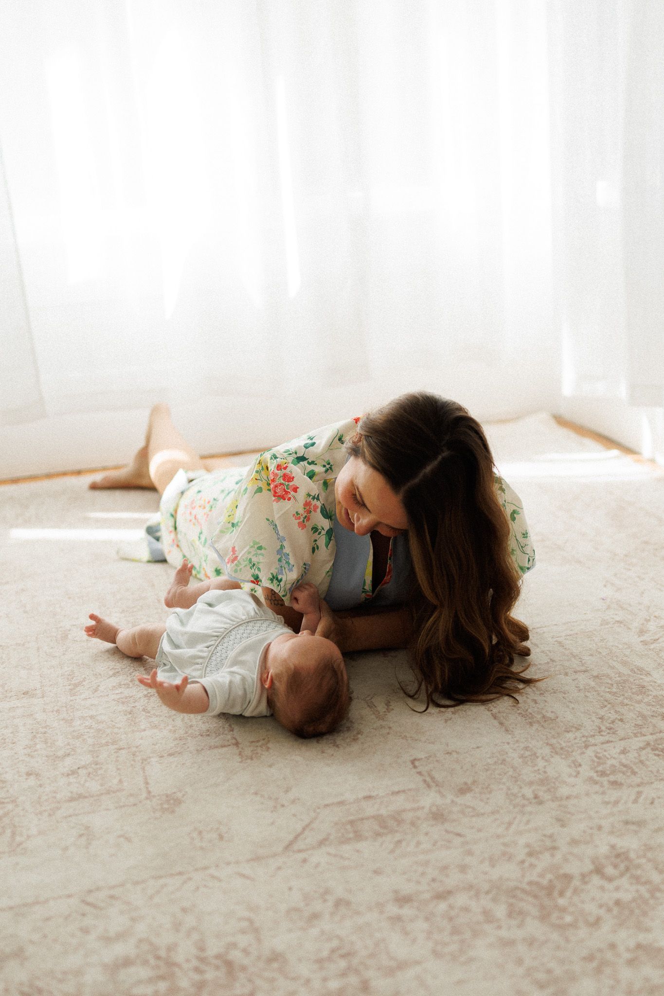 A smiling mother in a floral dress leans over her newborn baby on a soft carpet in a bright, sunlit room.
