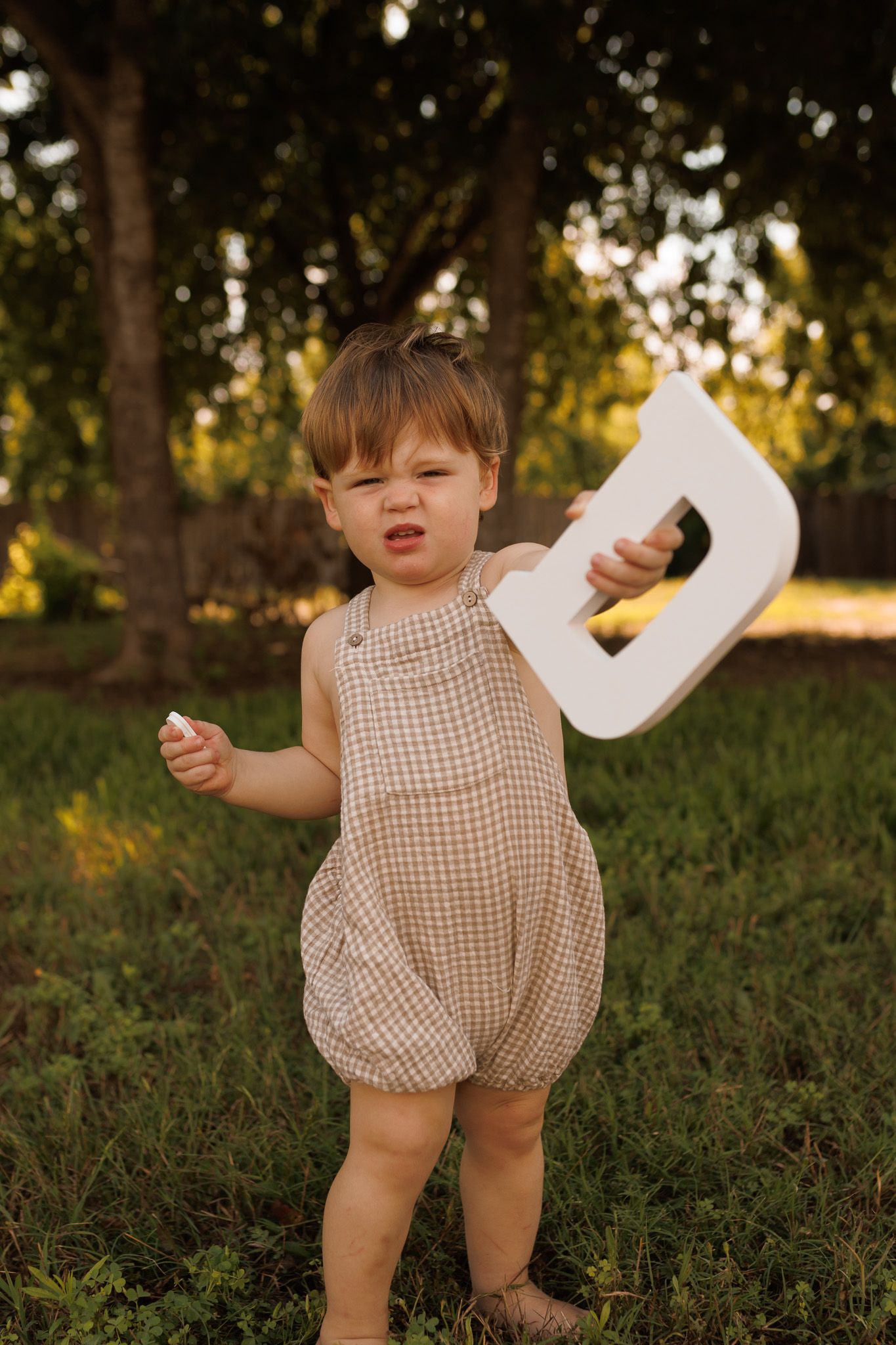 Small child in beige gingham romper plays outdoors at sunset while holding a white paper letter.