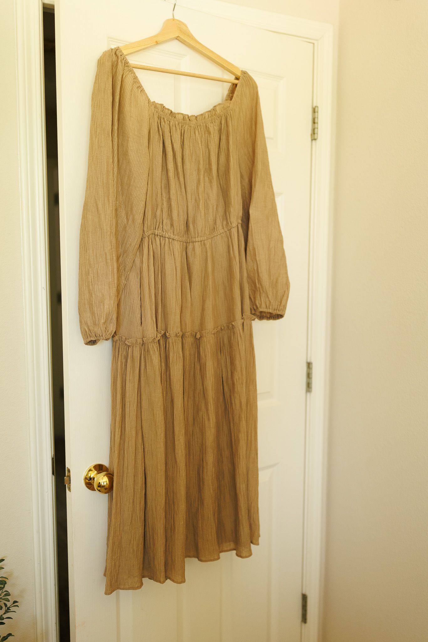 A tan bohemian-style maxi dress with long sleeves and tiered skirt hangs on a wooden hanger against a white door.