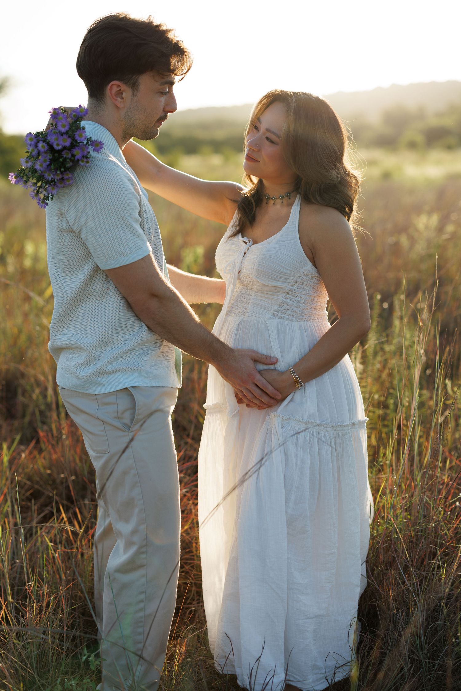 Couple in white outfits sharing a romantic moment in a sunlit field, woman in flowing dress holding flowers.