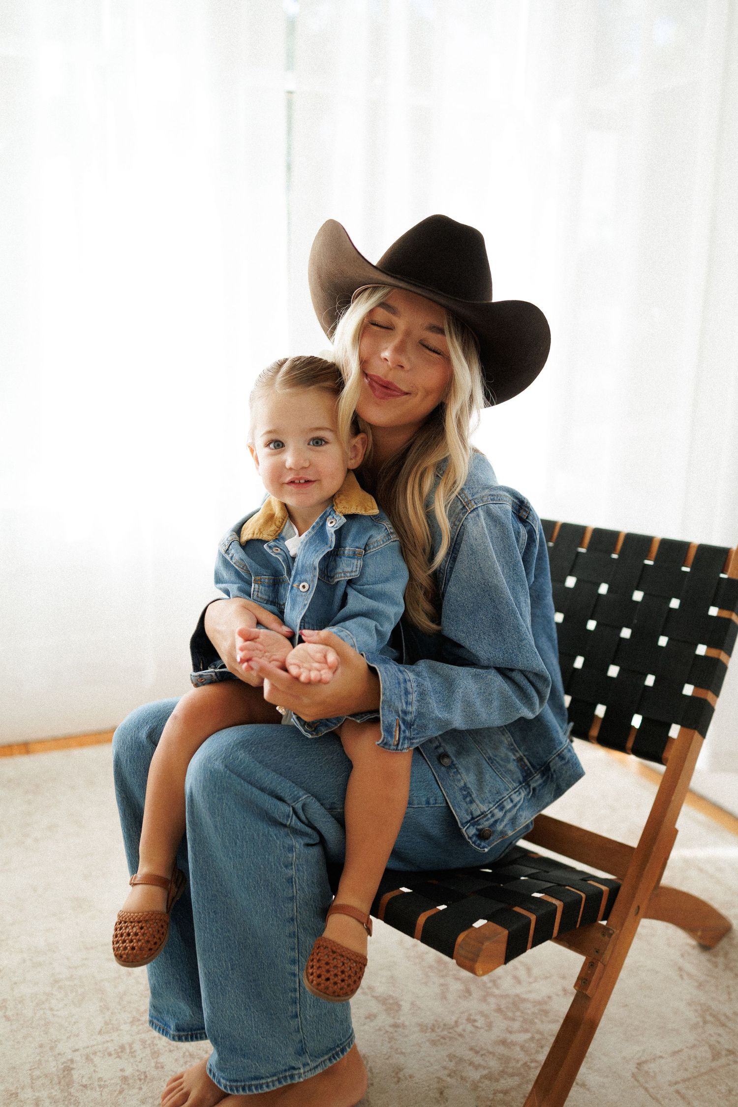 A blonde woman in a black cowboy hat and denim jacket holds a toddler in matching denim while sitting on a woven chair.