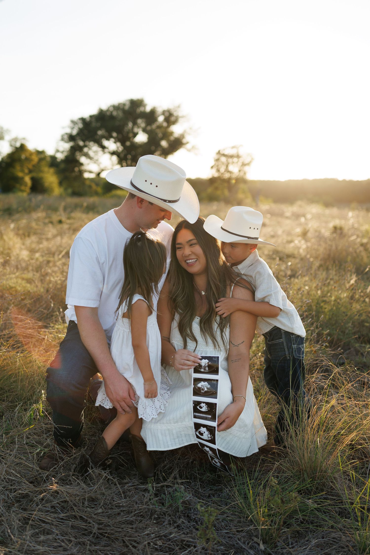 Family in white outfits and cowboy hats holding ultrasound photos in a golden field at sunset.