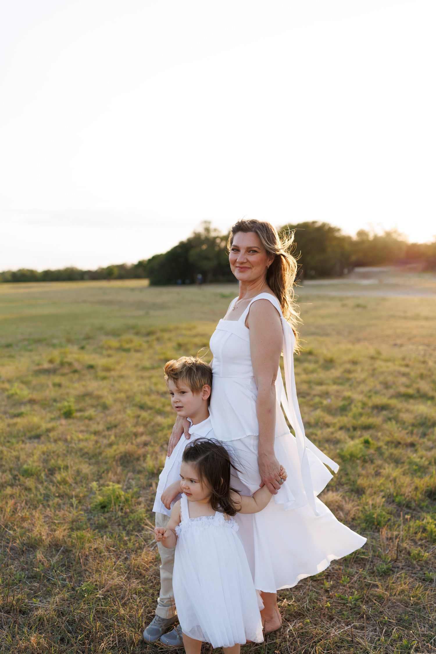 A mother and two children in white dresses stand in a sunlit field at golden hour.