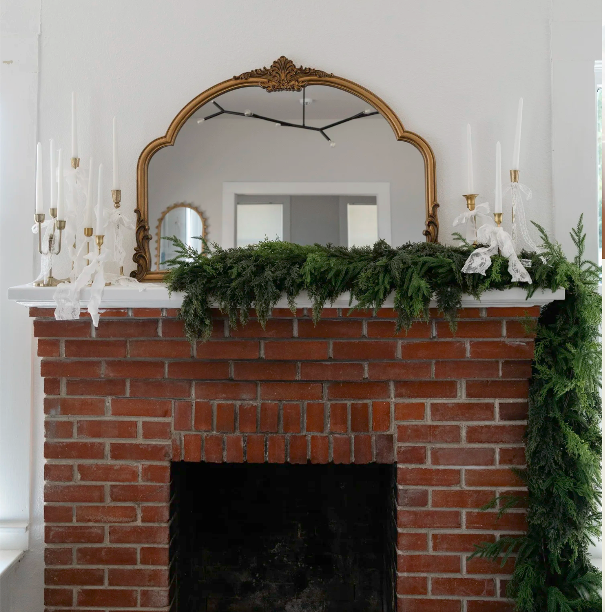 Red brick fireplace with gold arched mirror, evergreen garland, white candles, and holiday decor on white mantel.