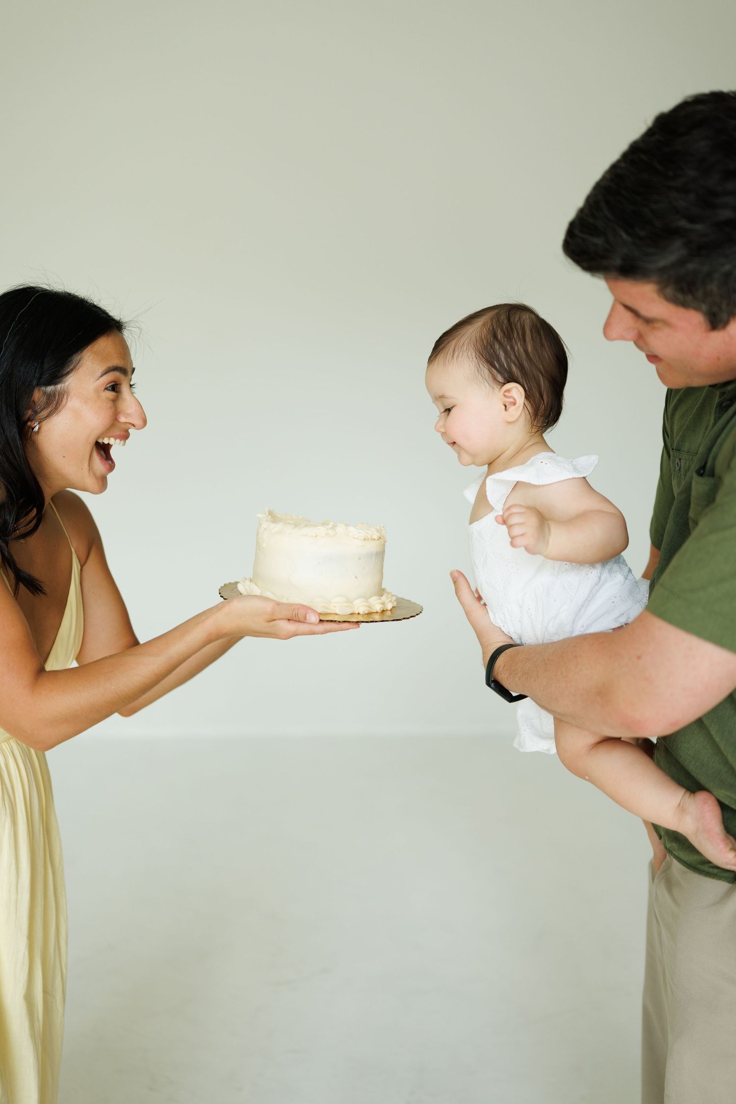 A woman presents a white birthday cake to a baby held by a man during a first birthday smash cake photoshoot.