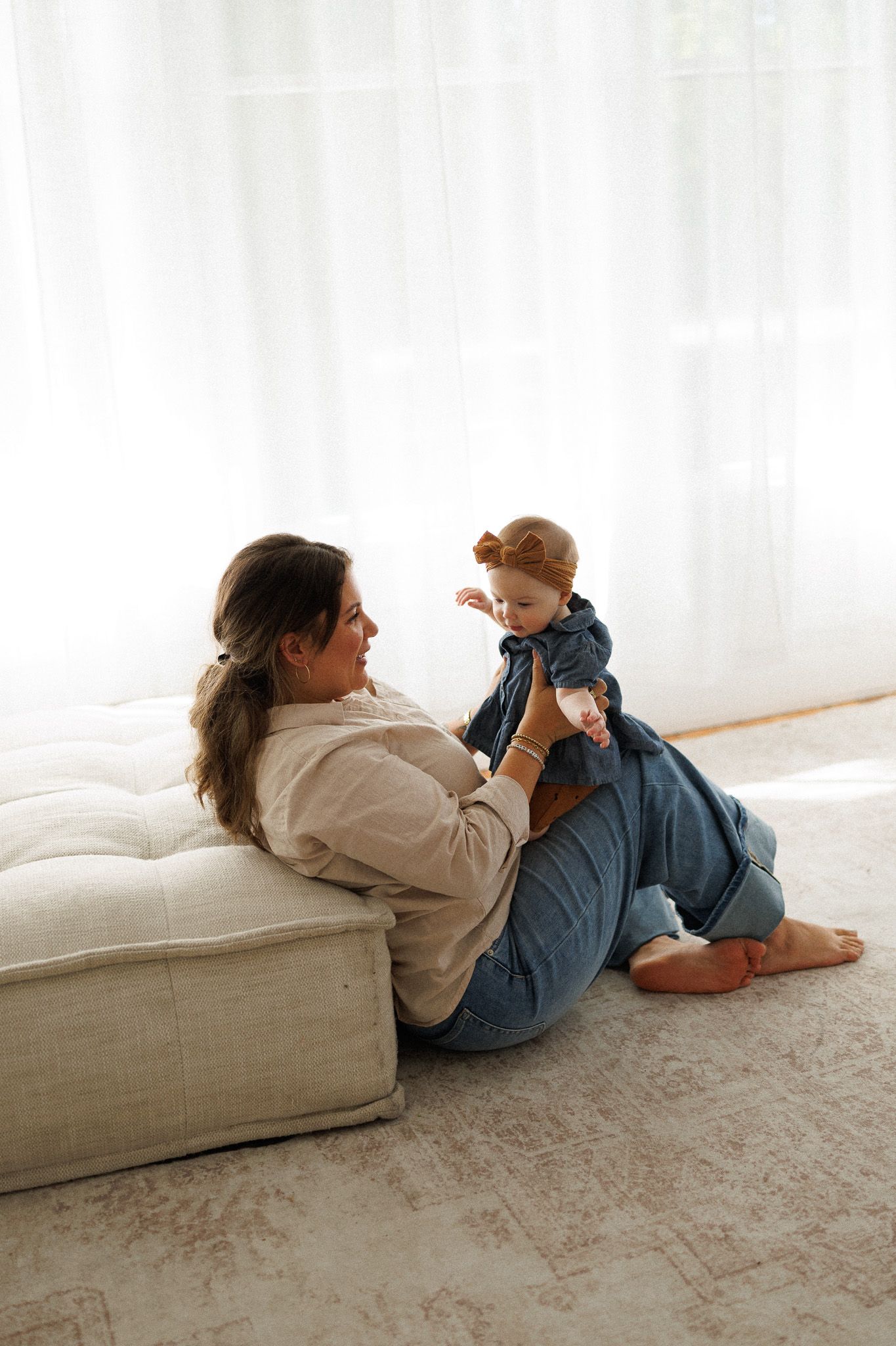 Mother in casual outfit sits on floor playing with her baby in a bright, airy living room.