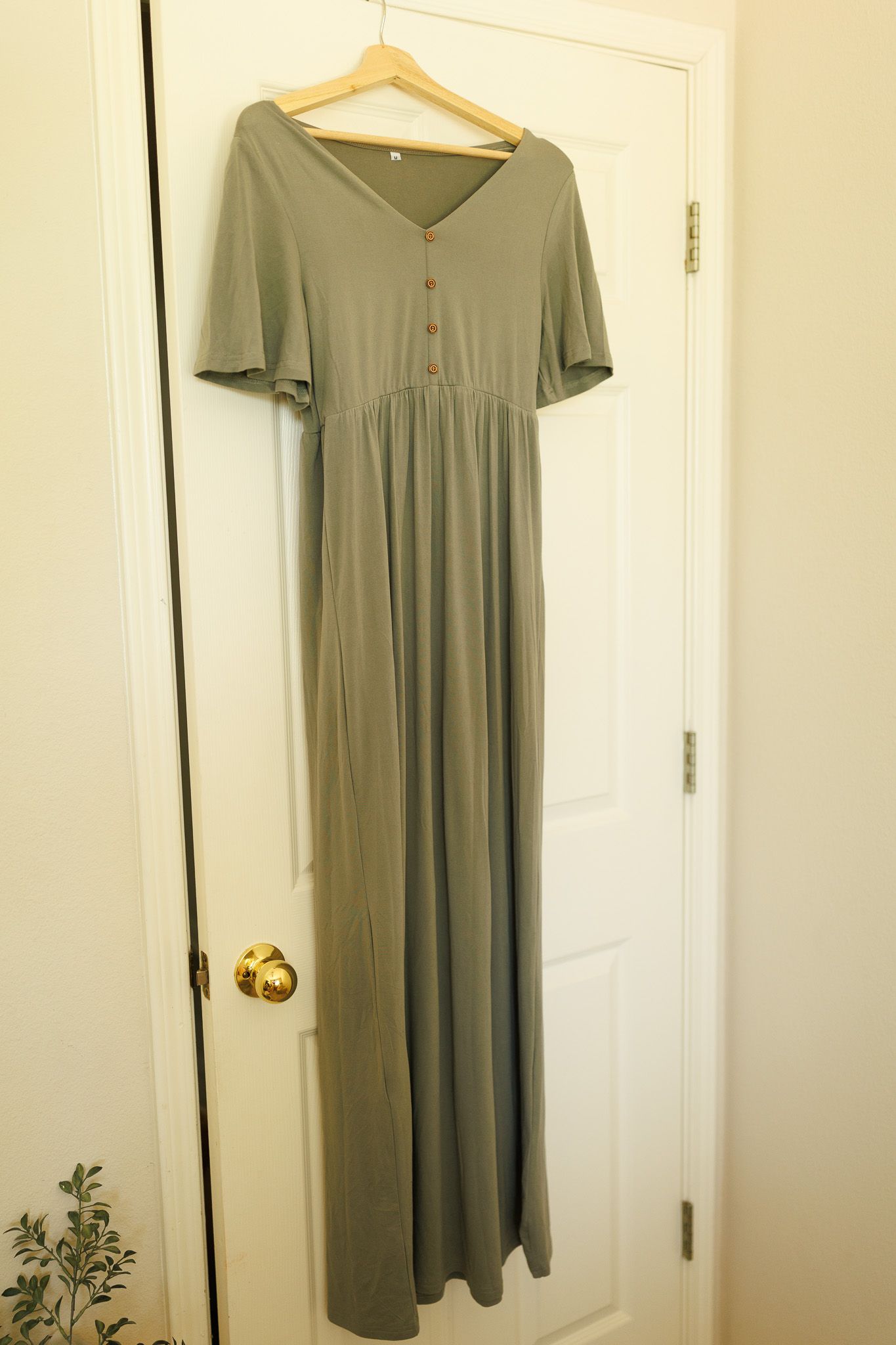 A sage green maxi dress with short flutter sleeves and button details hangs on a wooden hanger against a white door.