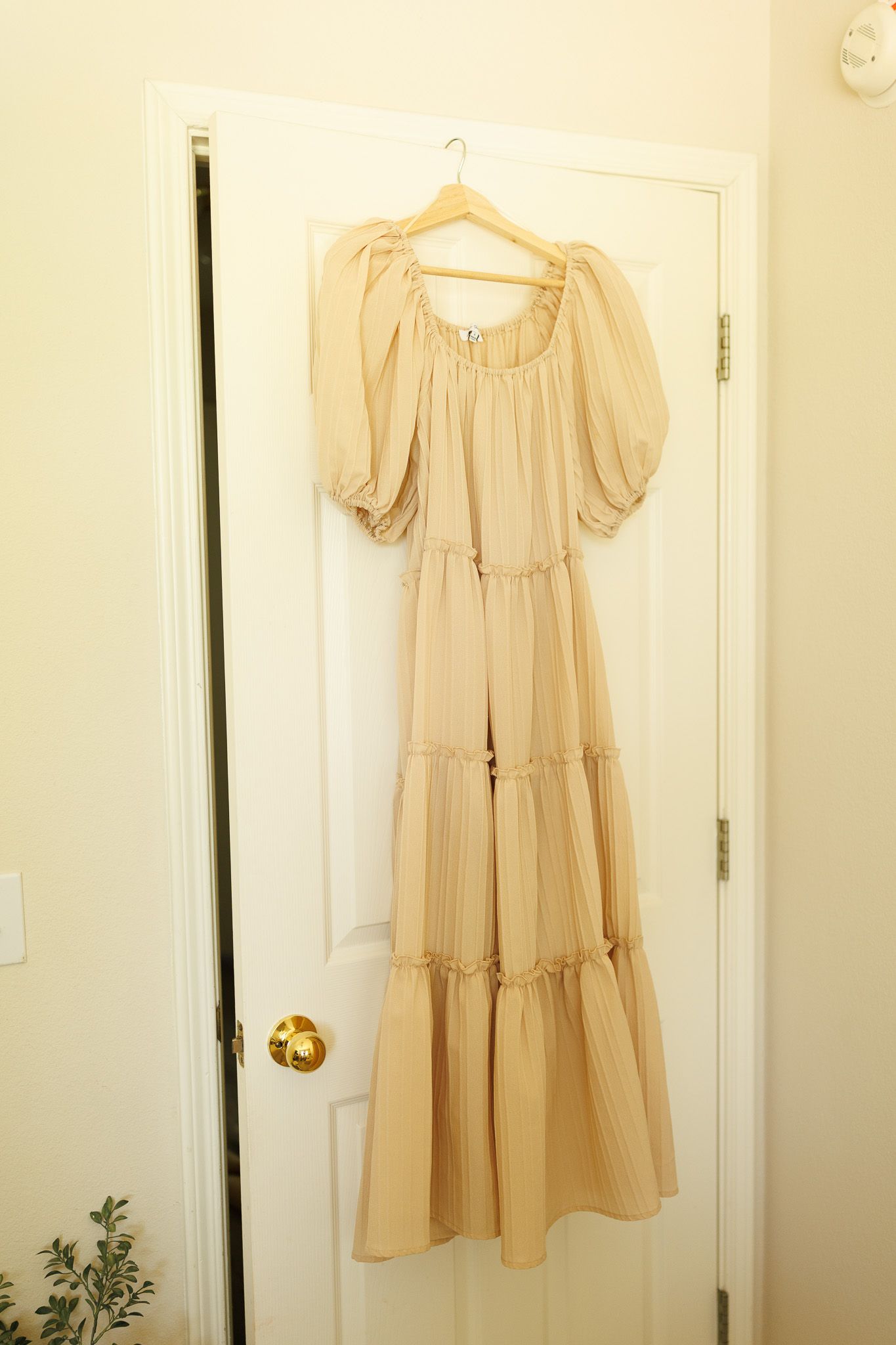A beige peasant-style maxi dress with tiered ruffles and puffed sleeves hanging on a wooden hanger against a white door.