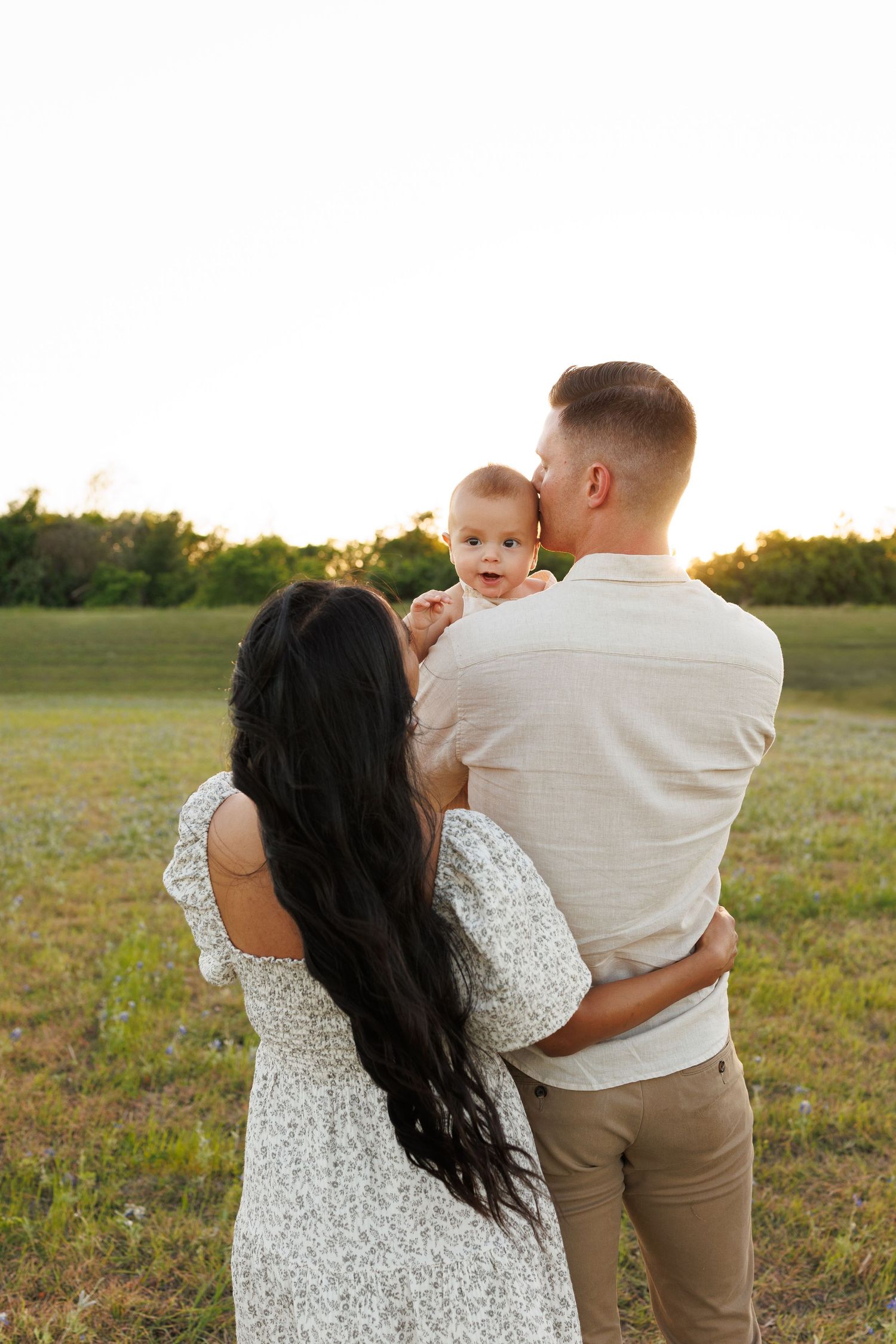 Family shares a tender moment together in a sunlit field at golden hour wearing coordinated white and neutral outfits.