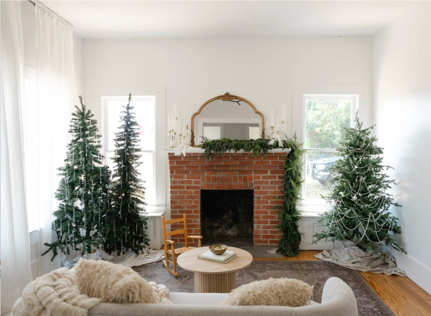 Cozy living room with brick fireplace, gold mirror, and three Christmas trees flanking the mantel.