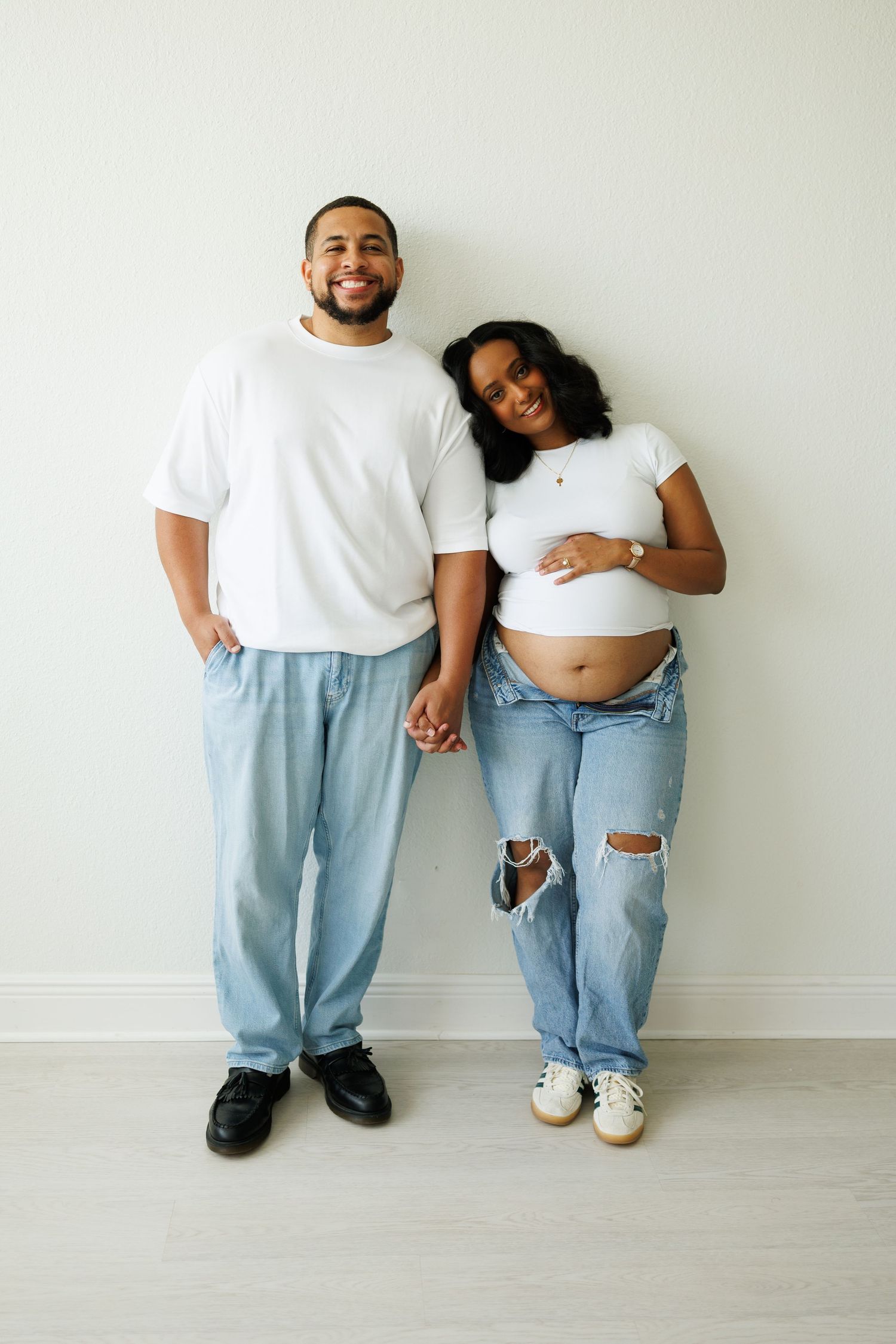A smiling couple in matching white tees and jeans pose together, the woman proudly showing her baby bump.
