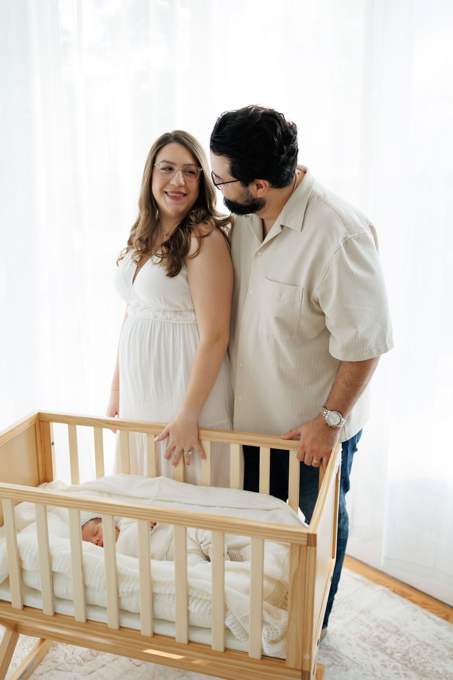 A couple in white clothing stands beside a wooden baby crib in a bright nursery room.