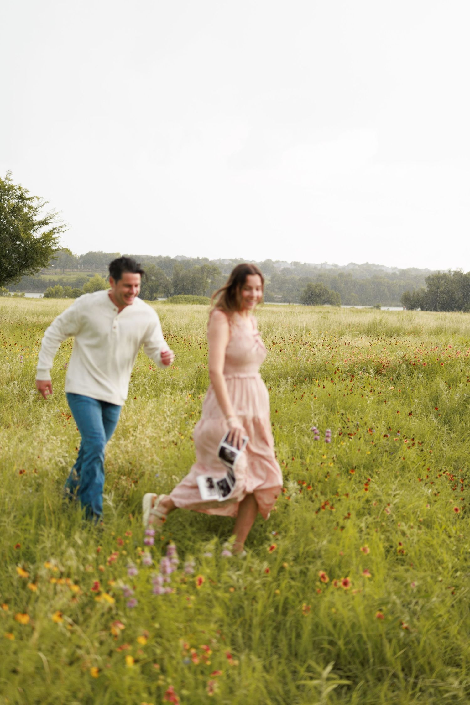 A happy couple walks through a wildflower meadow, woman in pink dress holding ultrasound photos, man in white shirt and jeans.