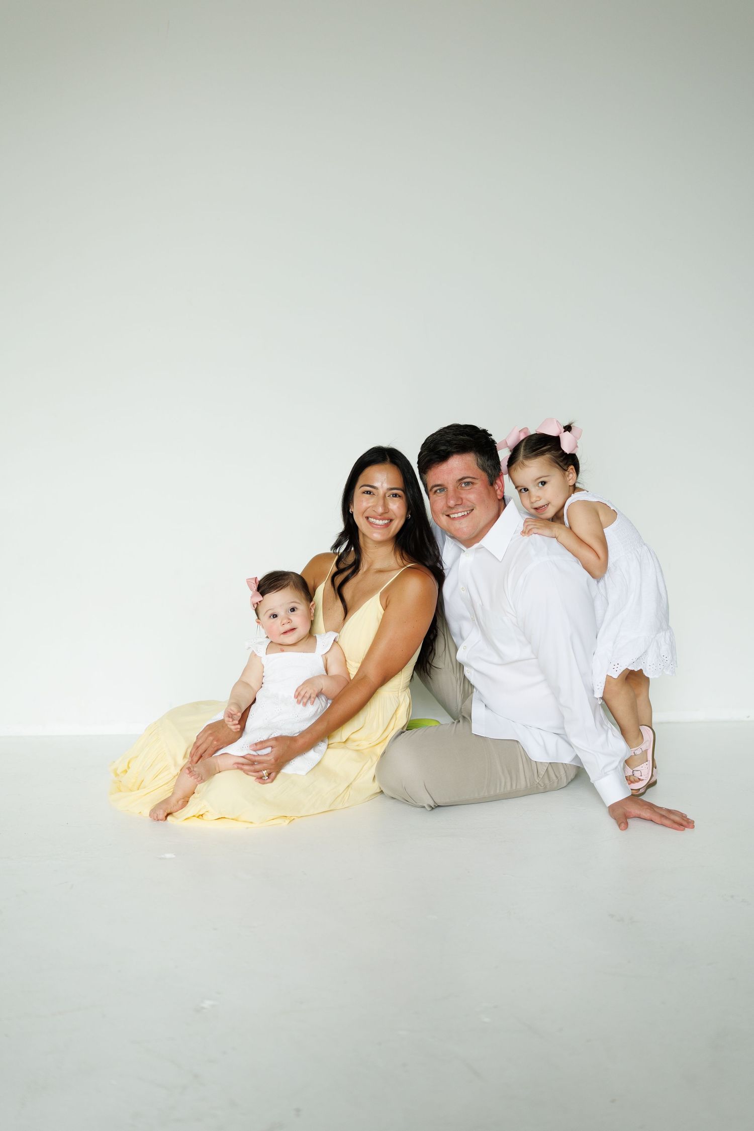 Happy family of four sitting on white studio floor, parents holding two young daughters in yellow and white outfits.